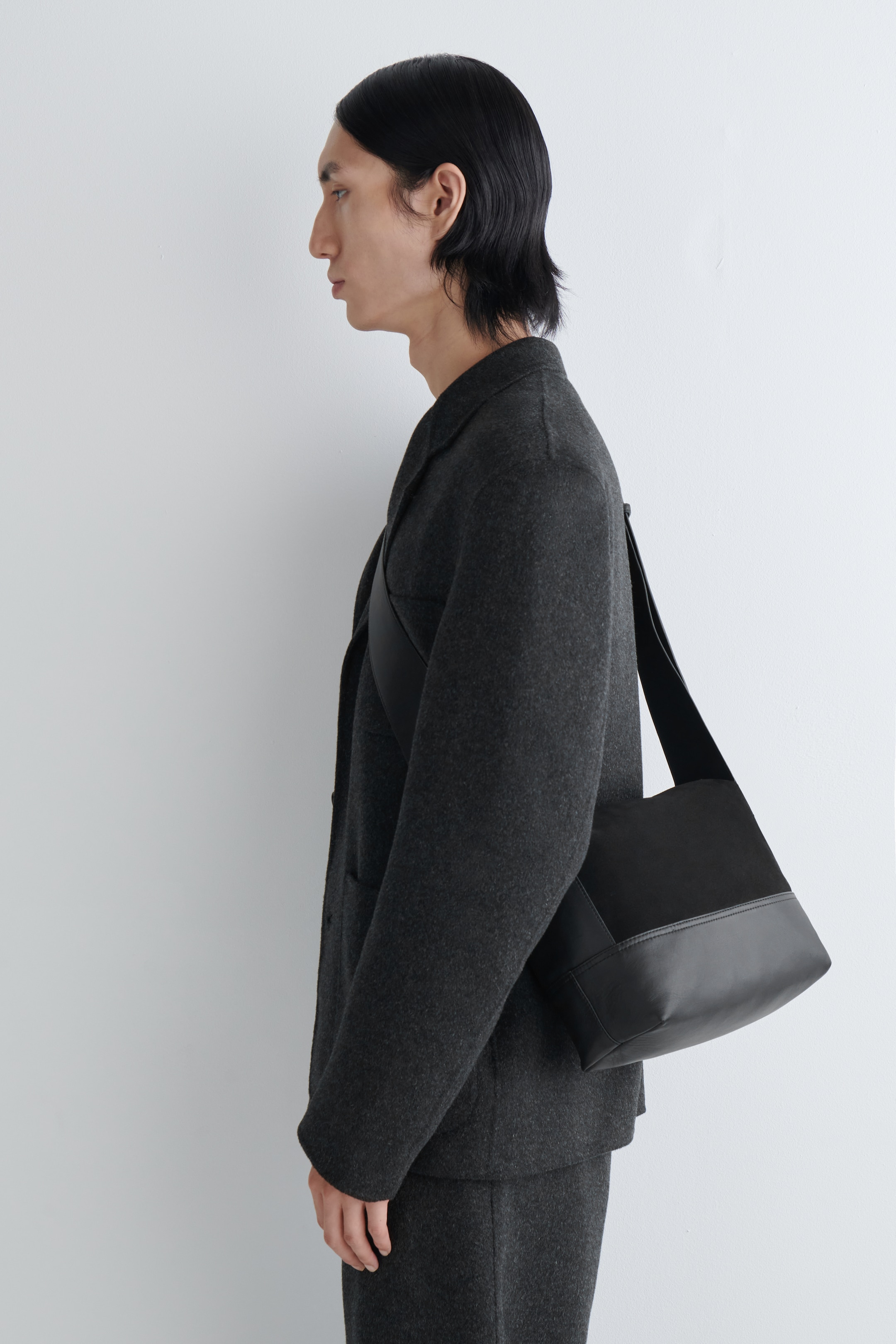 Agrandir l'image: Un sac noir porté en bandoulière est incliné pour montrer son profil latéral structuré. Le sac présente une large sangle plate et un design à deux matériaux, avec une section supérieure en tissu noir mat et un panneau inférieur en cuir lisse formant sa base.
