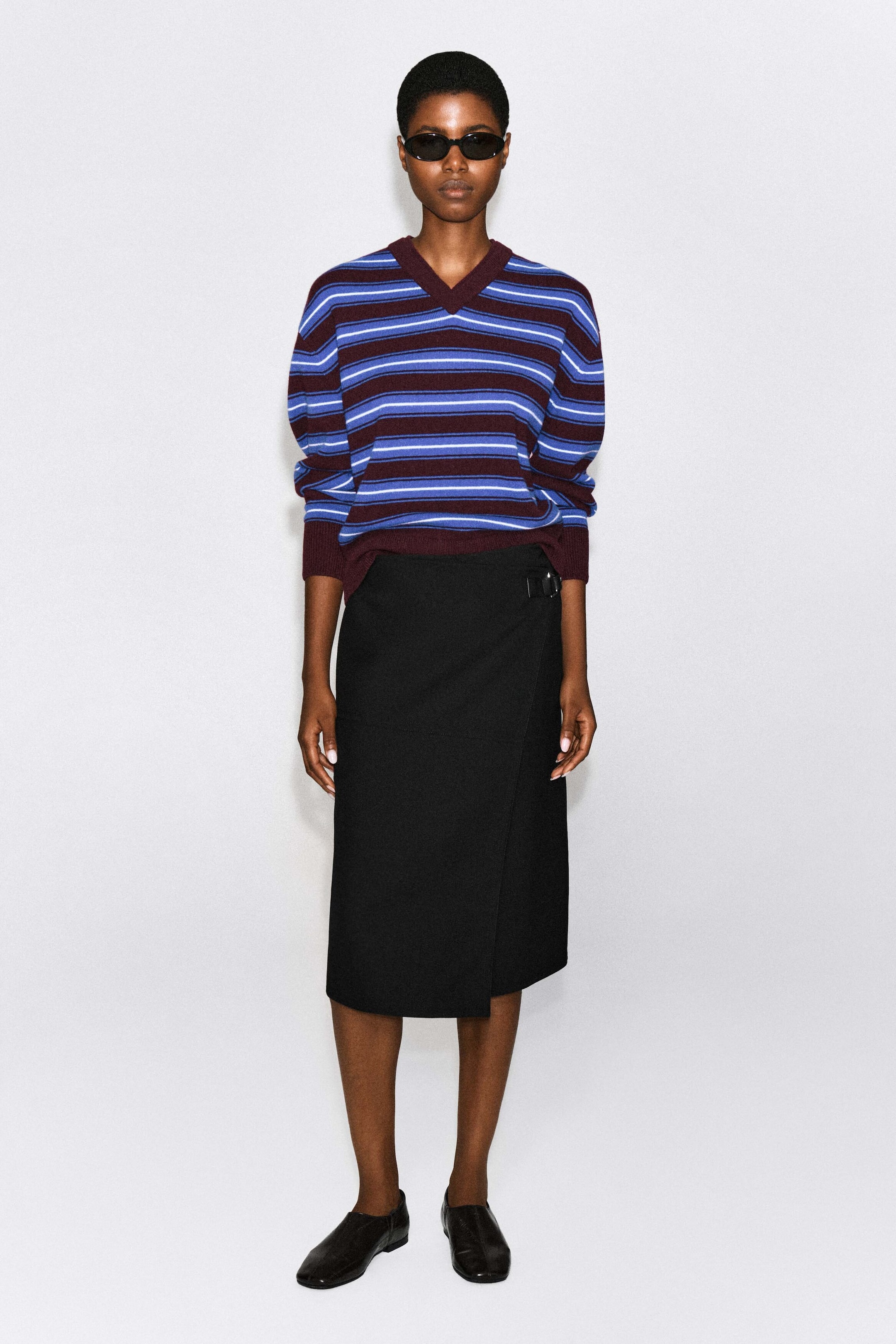 The merino wool layered V-neck sweater, featuring horizontal stripes in blue, white, and burgundy, is worn by the model. The sweater is styled with a black midi wrap skirt, black sunglasses, and black slip-on shoes, highlighting its relaxed fit, ribbed burgundy cuffs, and double-layered V-neckline. The merino wool layered V-neck sweater, featuring horizontal stripes in blue, white, and burgundy, is worn by the model. The sweater is styled with a black midi wrap skirt, black sunglasses, and black slip-on shoes, highlighting its relaxed fit, ribbed burgundy cuffs, and double-layered V-neckline.