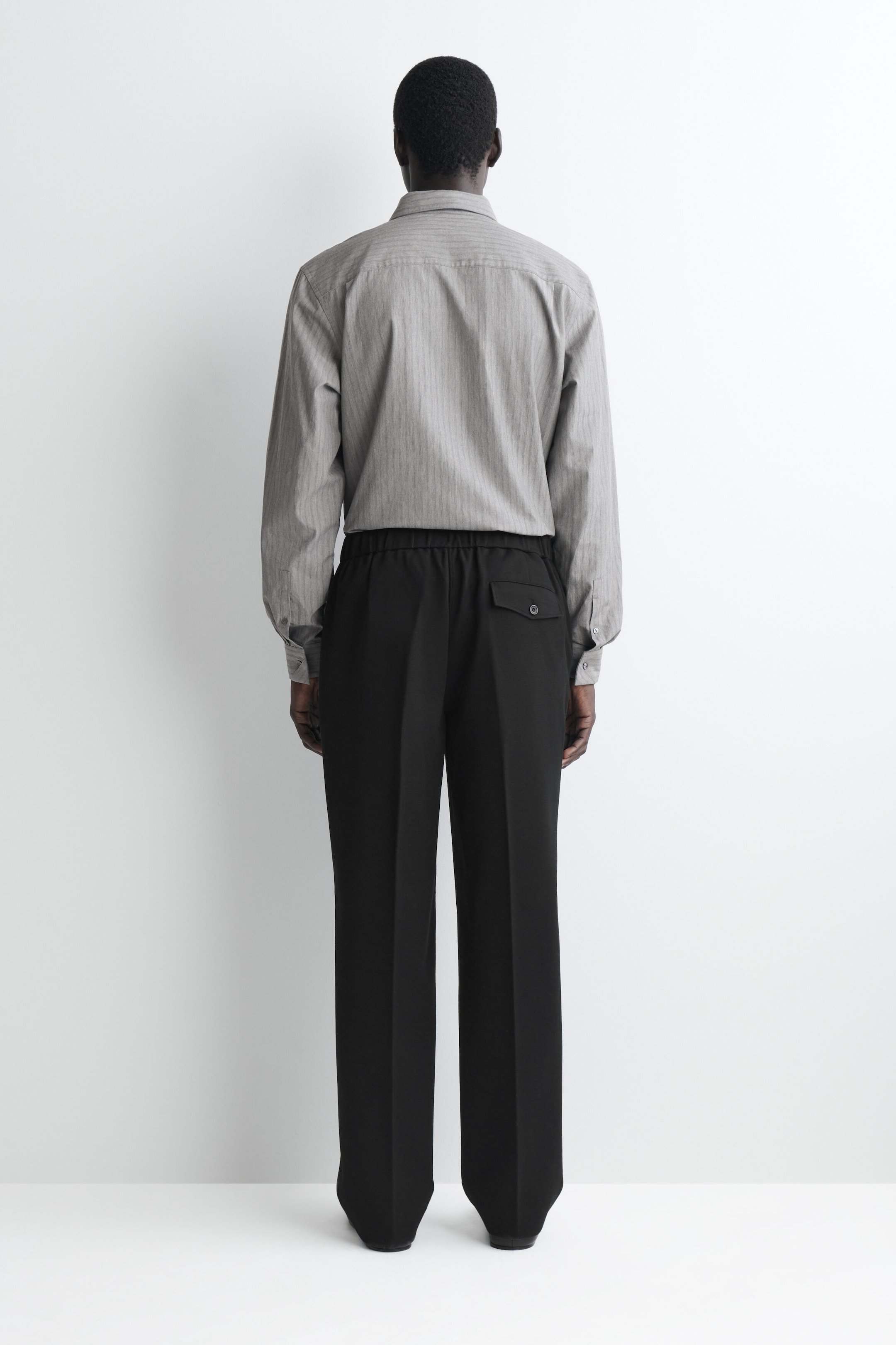 These black trousers, shown from the back, feature an elastic waistband, sharp center creases, and a single buttoned flap pocket on the right side. They are styled with a long-sleeved, finely textured gray button-down shirt tucked neatly into the waistband. These black trousers, shown from the back, feature an elastic waistband, sharp center creases, and a single buttoned flap pocket on the right side. They are styled with a long-sleeved, finely textured gray button-down shirt tucked neatly into the waistband.