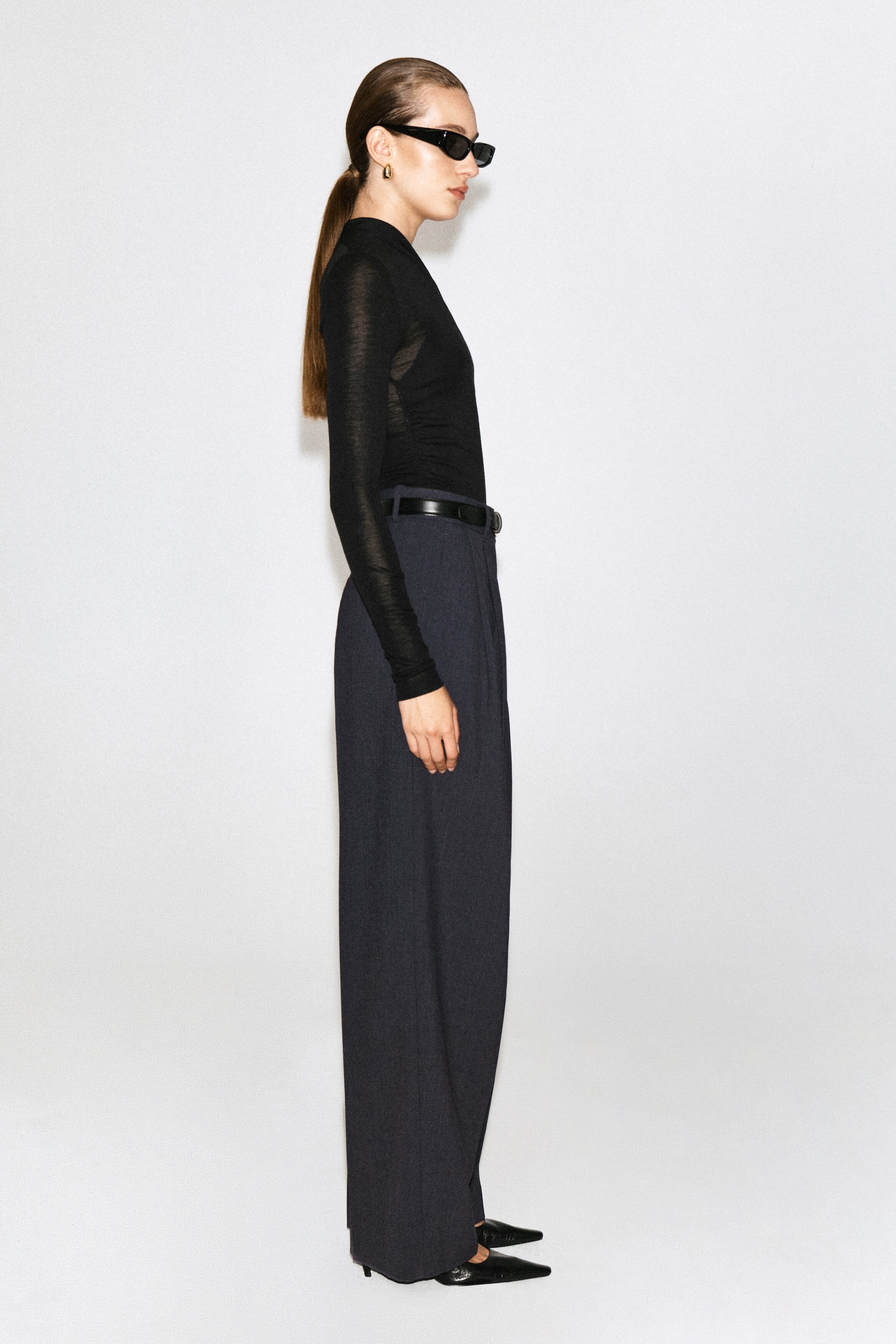 A black, slim-fit, long-sleeved top is being worn by the model, who is standing in profile. The fine-knit top features a high collar and is tucked into dark, wide-leg trousers styled with a black belt. A black, slim-fit, long-sleeved top is being worn by the model, who is standing in profile. The fine-knit top features a high collar and is tucked into dark, wide-leg trousers styled with a black belt.