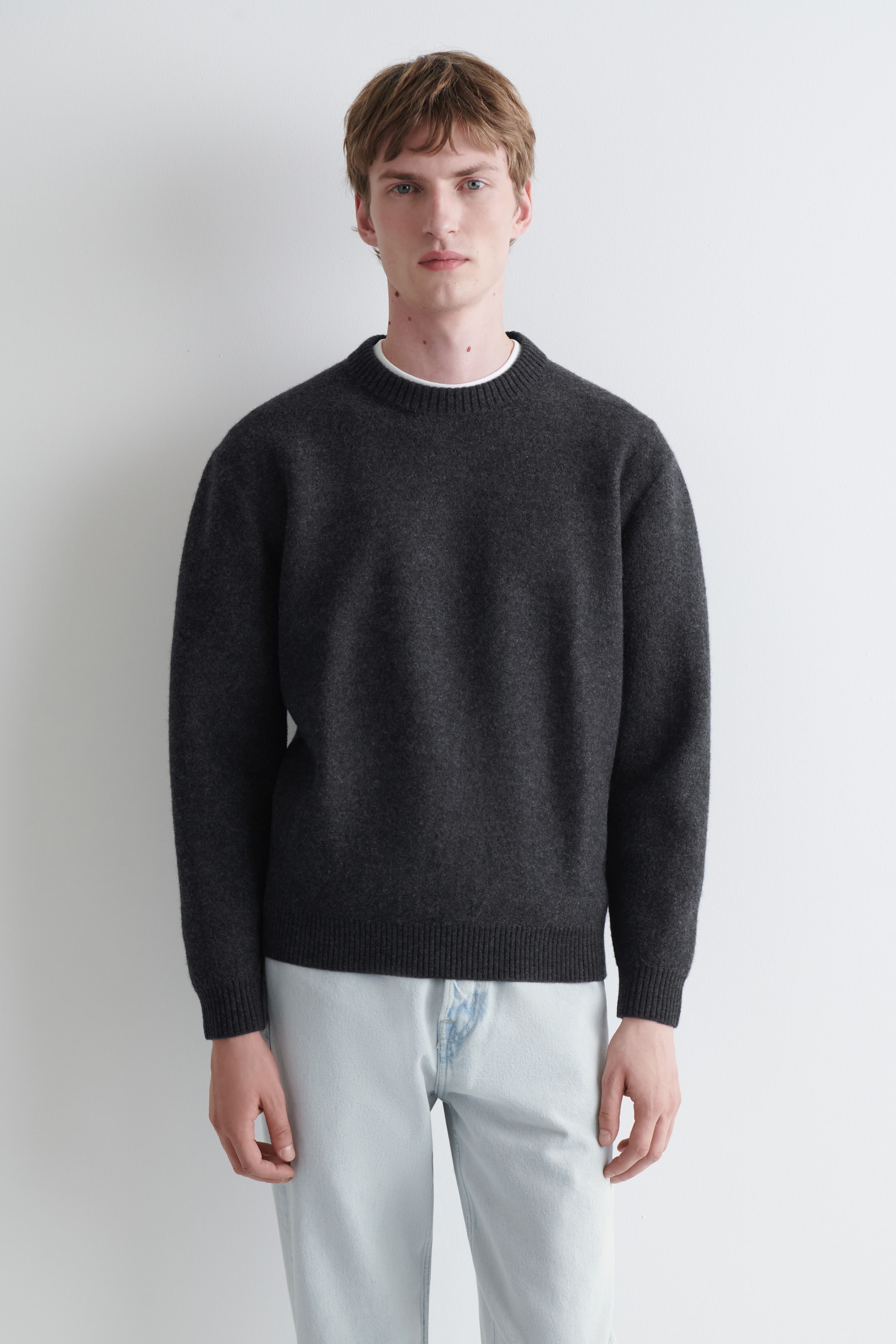 Grotere afbeelding bekijken: Het model draagt een donkergrijze trui met ronde hals. Deze heeft een geribde kraag, manchetten en zoom, en is gelaagd over een wit T-shirt.