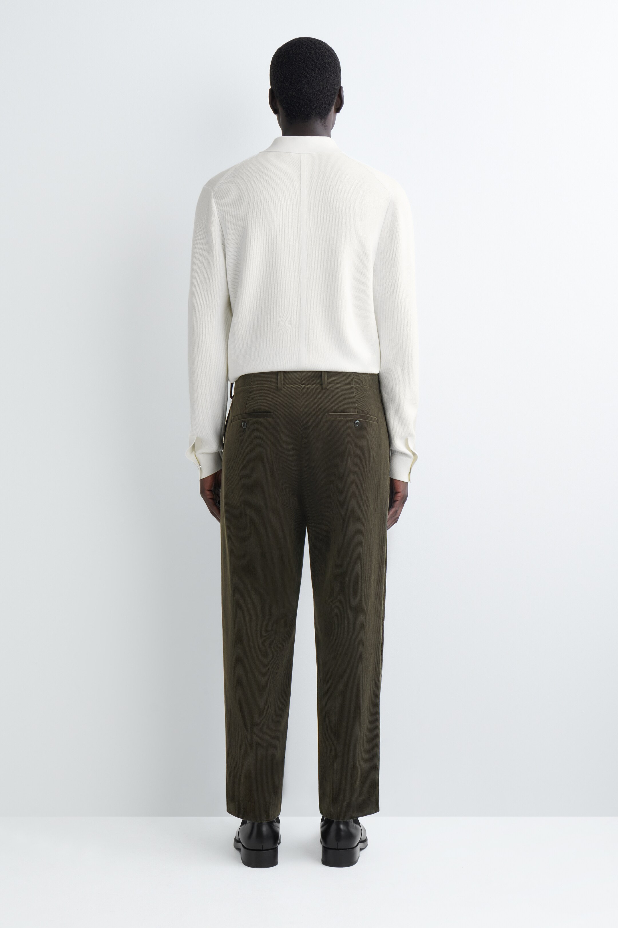 Agrandir l'image: Un pantalon en velours côtelé vert olive foncé est présenté de dos, avec une coupe droite et décontractée, et deux poches arrière boutonnées. Une chemise à manches longues blanc cassé avec un col officier et une couture verticale dans le dos est portée rentrée dans le pantalon.