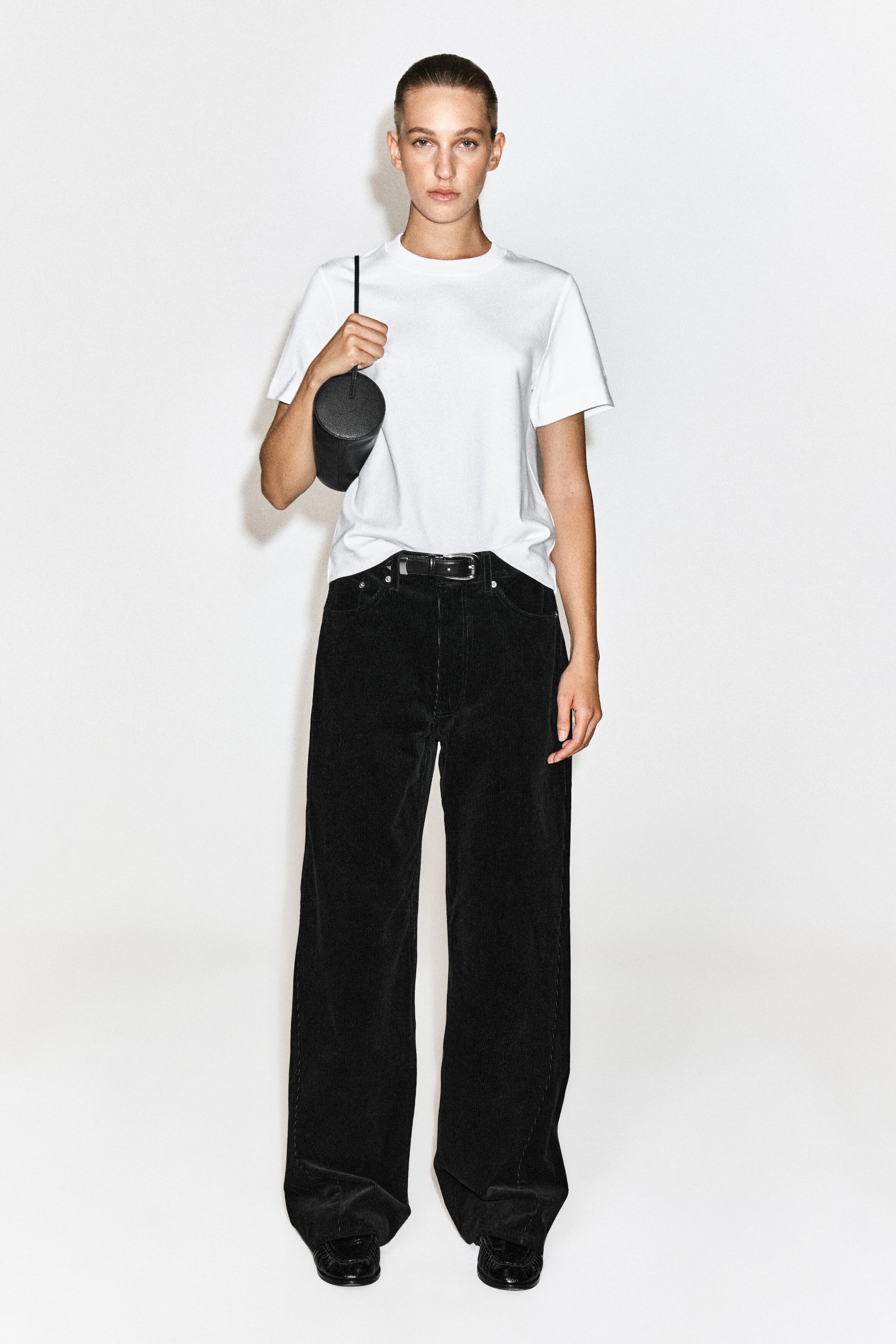 The model is wearing black wide-leg corduroy trousers, paired with a tucked-in white short-sleeve T-shirt and a black leather belt. The model is holding a small black cylindrical bag with a thin strap.