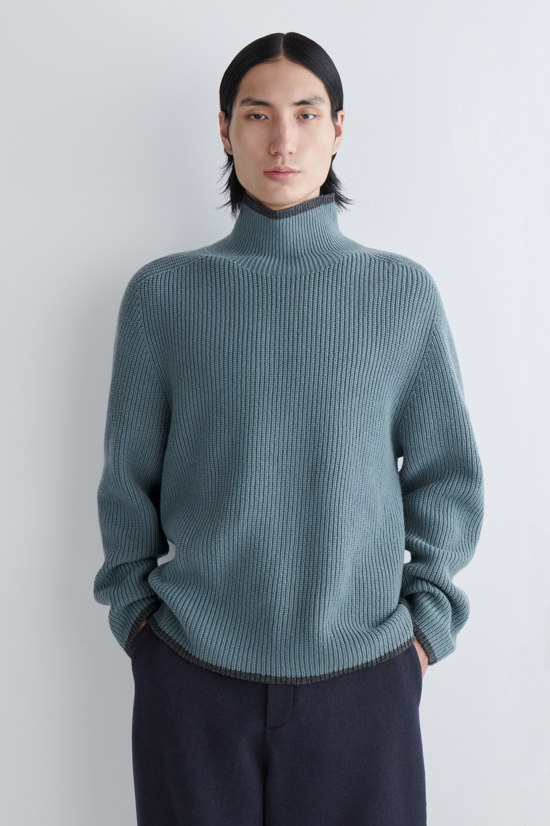 Agrandir l'image: Le pull à col roulé côtelé verticalement de couleur sarcelle est présenté de face, mettant en valeur sa coupe décontractée. Il présente une bordure contrastante gris foncé le long de son ourlet droit, qui est superposé à un pantalon bleu foncé.