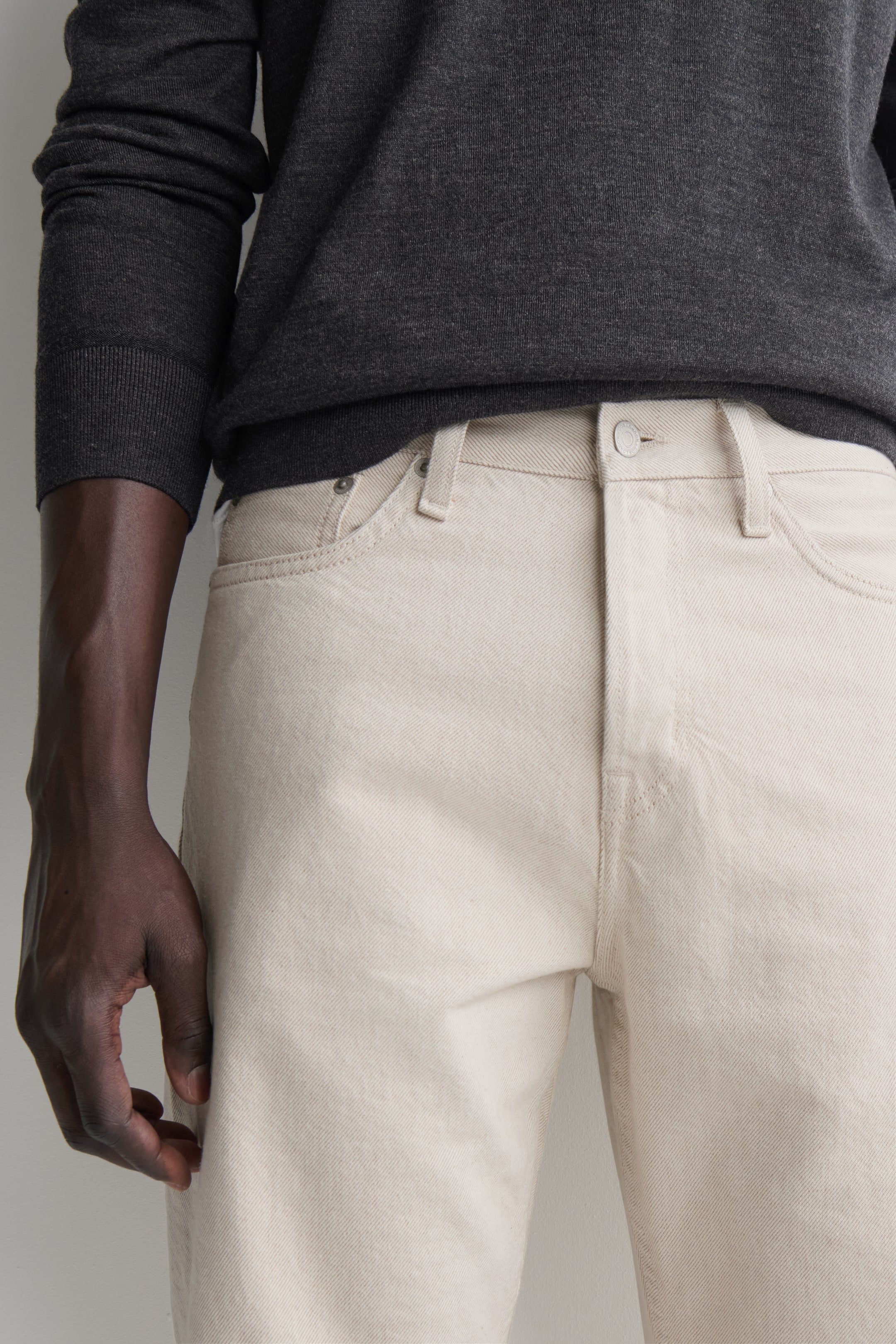 Visualizza immagine più grande: Questi jeans in denim bianco sporco presentano un classico design a cinque tasche ripreso da una prospettiva frontale, con passanti per cintura e chiusura con bottone metallico. Un maglione in maglia a maniche lunghe grigio scuro è appoggiato leggermente sopra il cinturino, mettendo in risalto la vestibilità strutturata dei jeans.