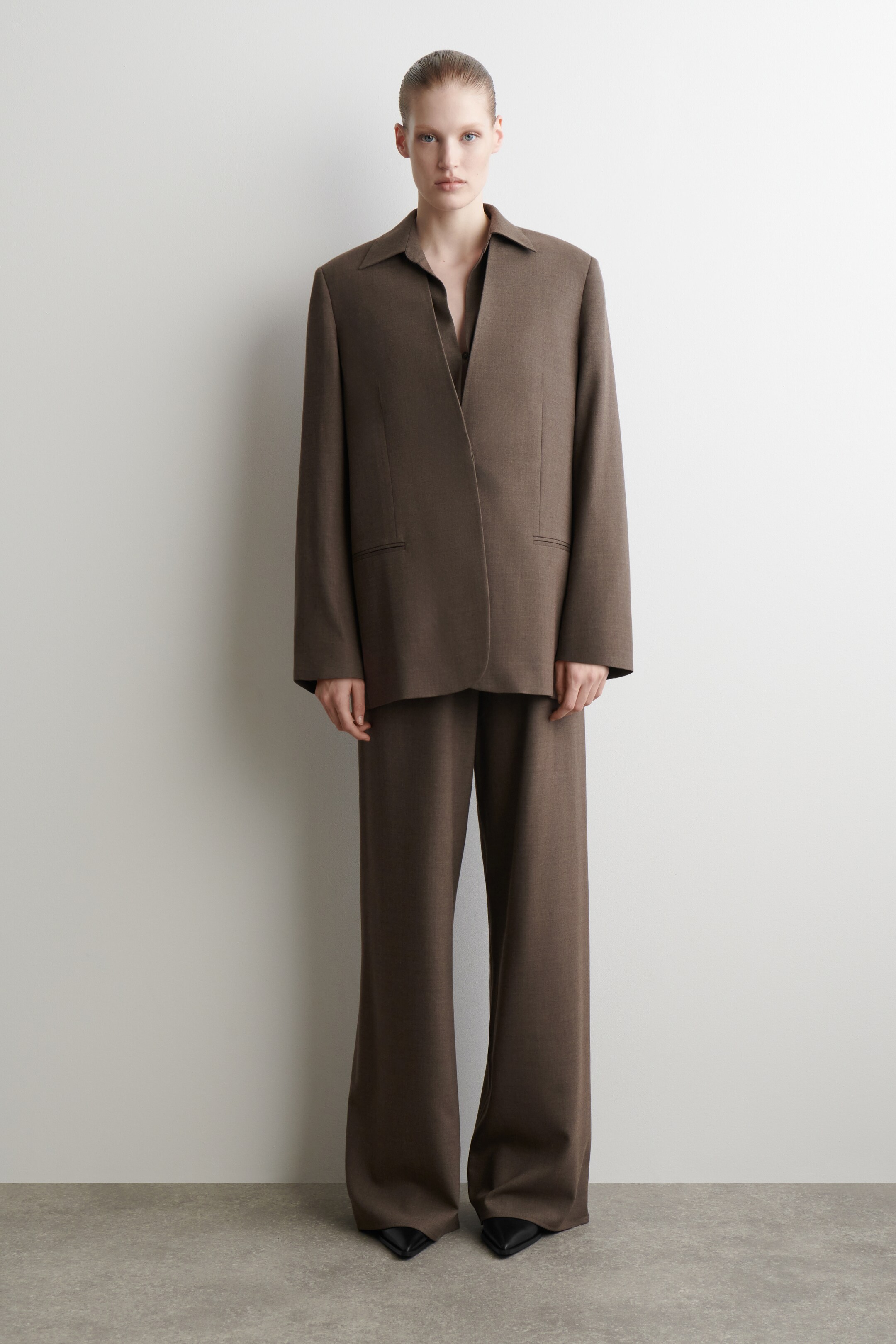 Agrandir l'image: Un costume marron, vu de face, présente un blazer surdimensionné à coupe décontractée avec des revers larges et souples et deux poches passepoilées horizontales. Le blazer est porté ouvert sur une chemise sombre à col. Un pantalon à jambes larges assorti complète le costume, tombant légèrement au-dessus de chaussures noires à bout pointu.