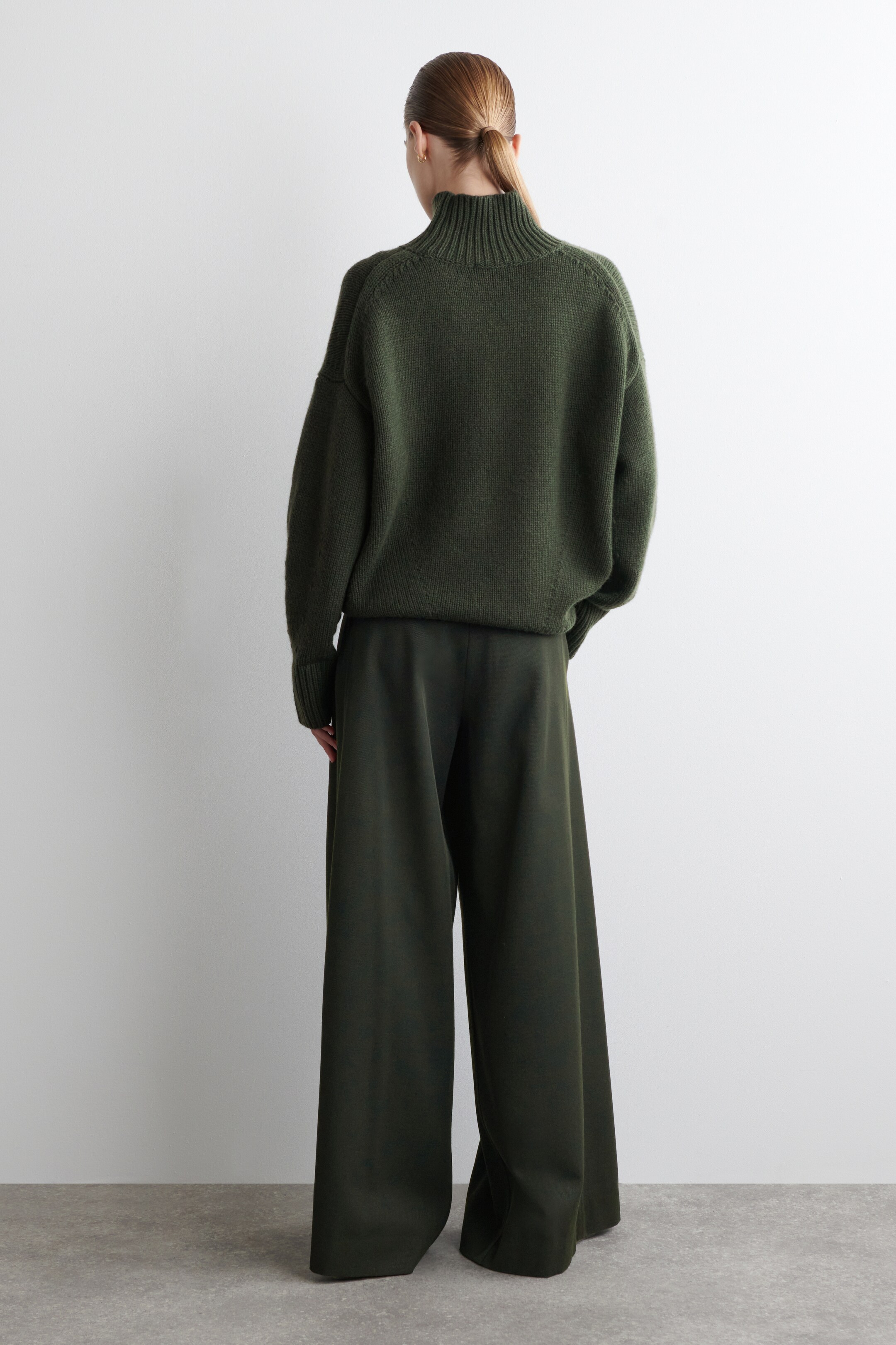 Visualizza immagine più grande: Un maglione lavorato a maglia oversize verde scuro viene indossato con pantaloni a gamba larga coordinati, visto da dietro. Il maglione presenta un collo alto a coste e maniche raglan, mentre i pantaloni cadono morbidi fino al pavimento.