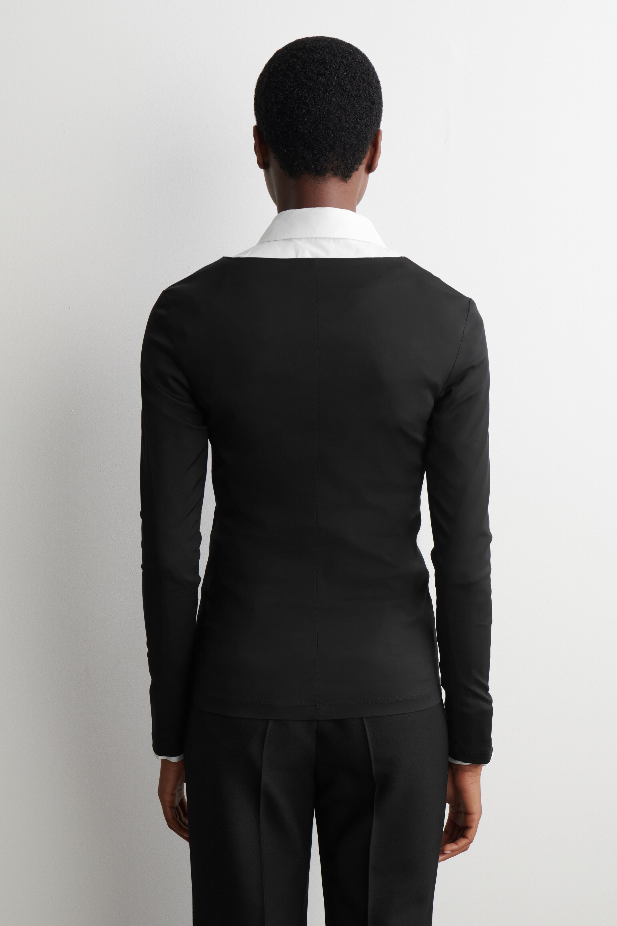 View larger image: A fitted black long-sleeved top is being viewed from the back, featuring a wide, shallow neckline that is revealing the collar of a white shirt underneath. A vertical seam is running down the center back of the top.