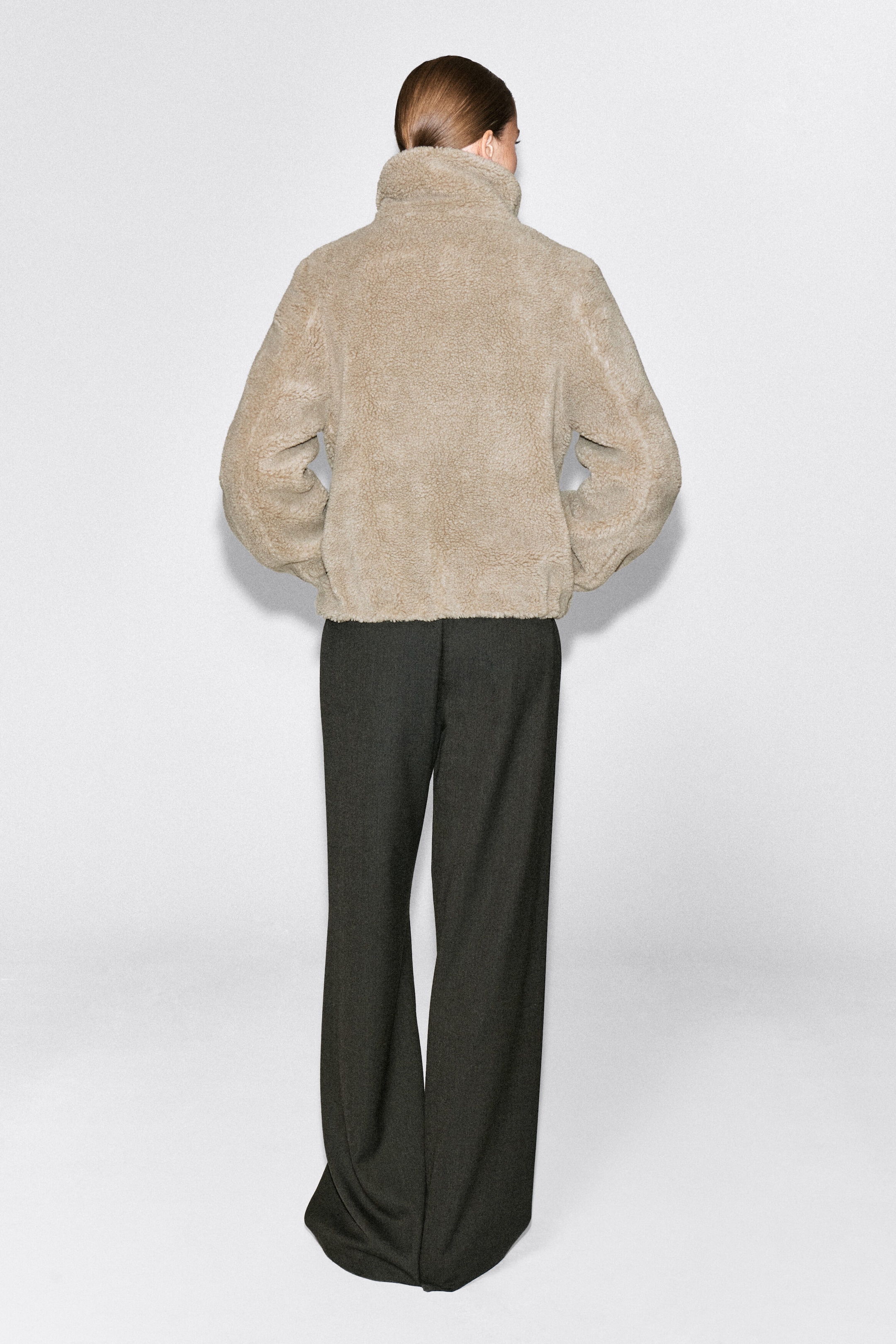 A beige funnel-neck teddy jacket with a textured, fleece-like finish is being worn by the model, shown from behind. The jacket features a high, standing collar and a relaxed fit, and is paired with dark, wide-leg trousers.
