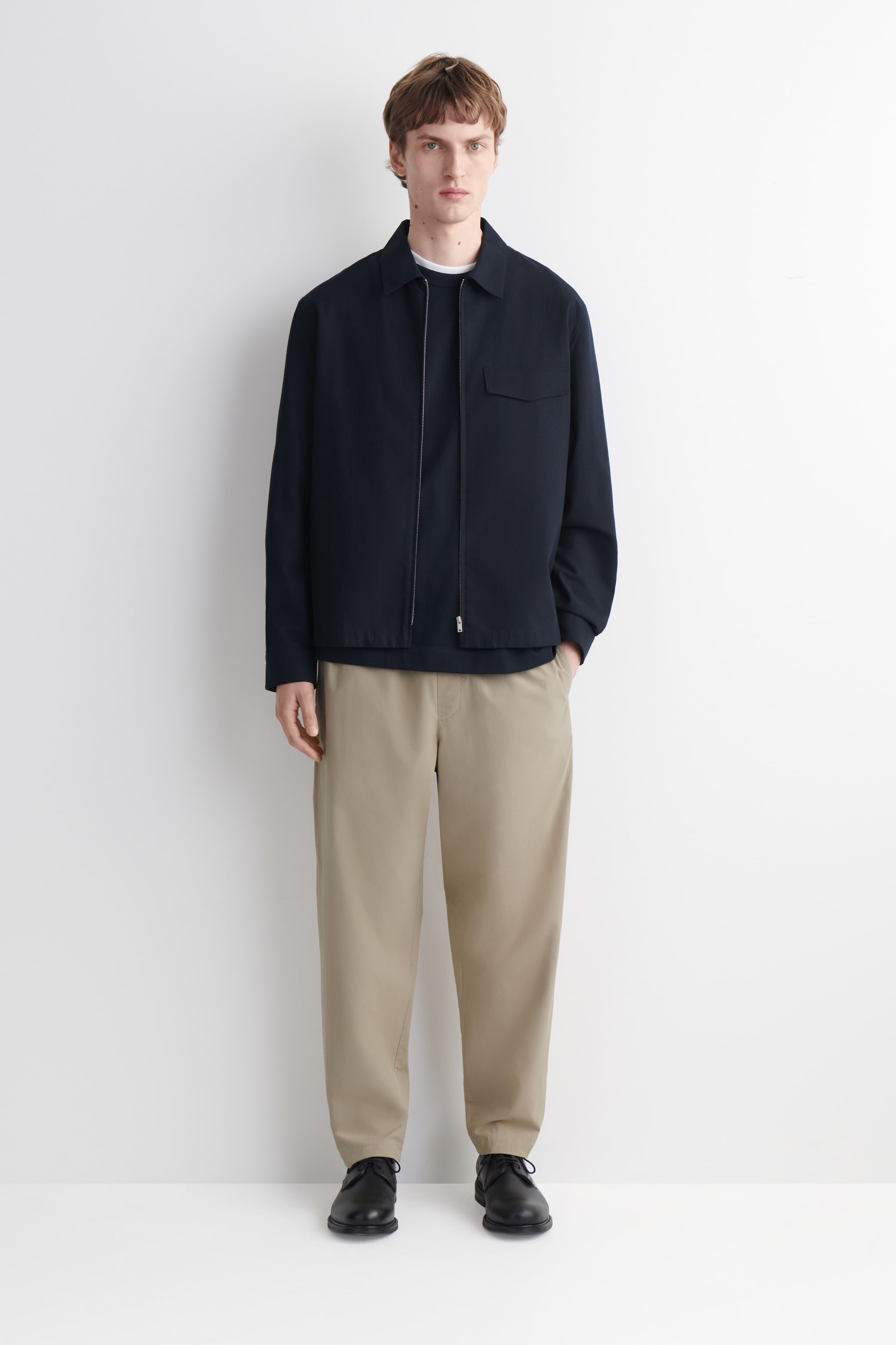 הצגת תמונה גדולה יותר: מעיל כחול כהה עם צווארון מוצג מזווית קדמית, כולל סגירת רוכסן כסף באורך מלא וכיס שטוח על החזה השמאלי. הוא מונח מעל חולצת טריקו לבנה עם צווארון עגול ובגד כחול כהה.