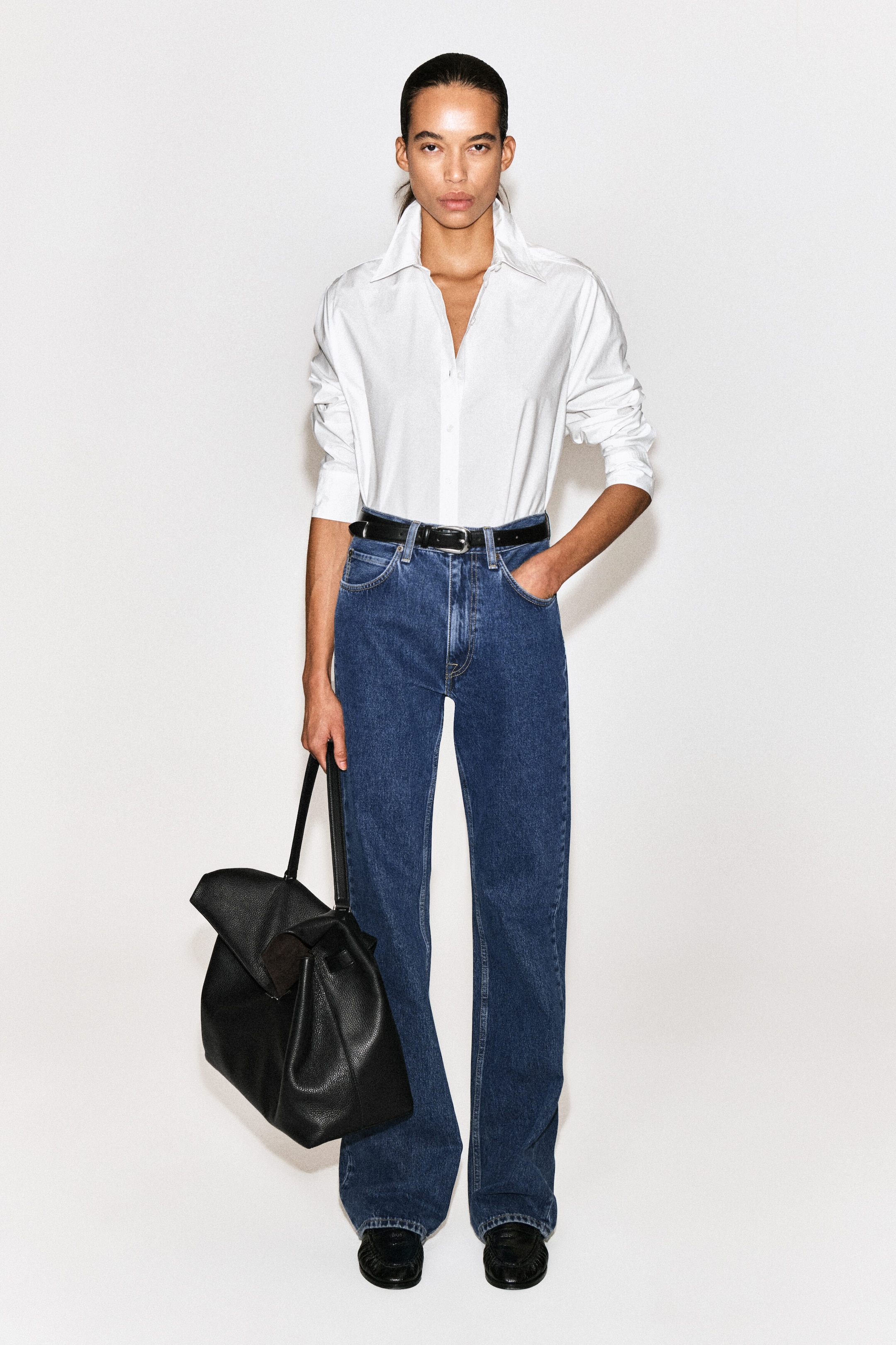 The classic cotton poplin shirt is shown from the front, featuring crisp white fabric, a pointed collar, and a button-down front. The shirt is tucked into high-waisted light blue jeans secured with a black belt, with the long sleeves pushed up to the forearms. The classic cotton poplin shirt is shown from the front, featuring crisp white fabric, a pointed collar, and a button-down front. The shirt is tucked into high-waisted light blue jeans secured with a black belt, with the long sleeves pushed up to the forearms.
