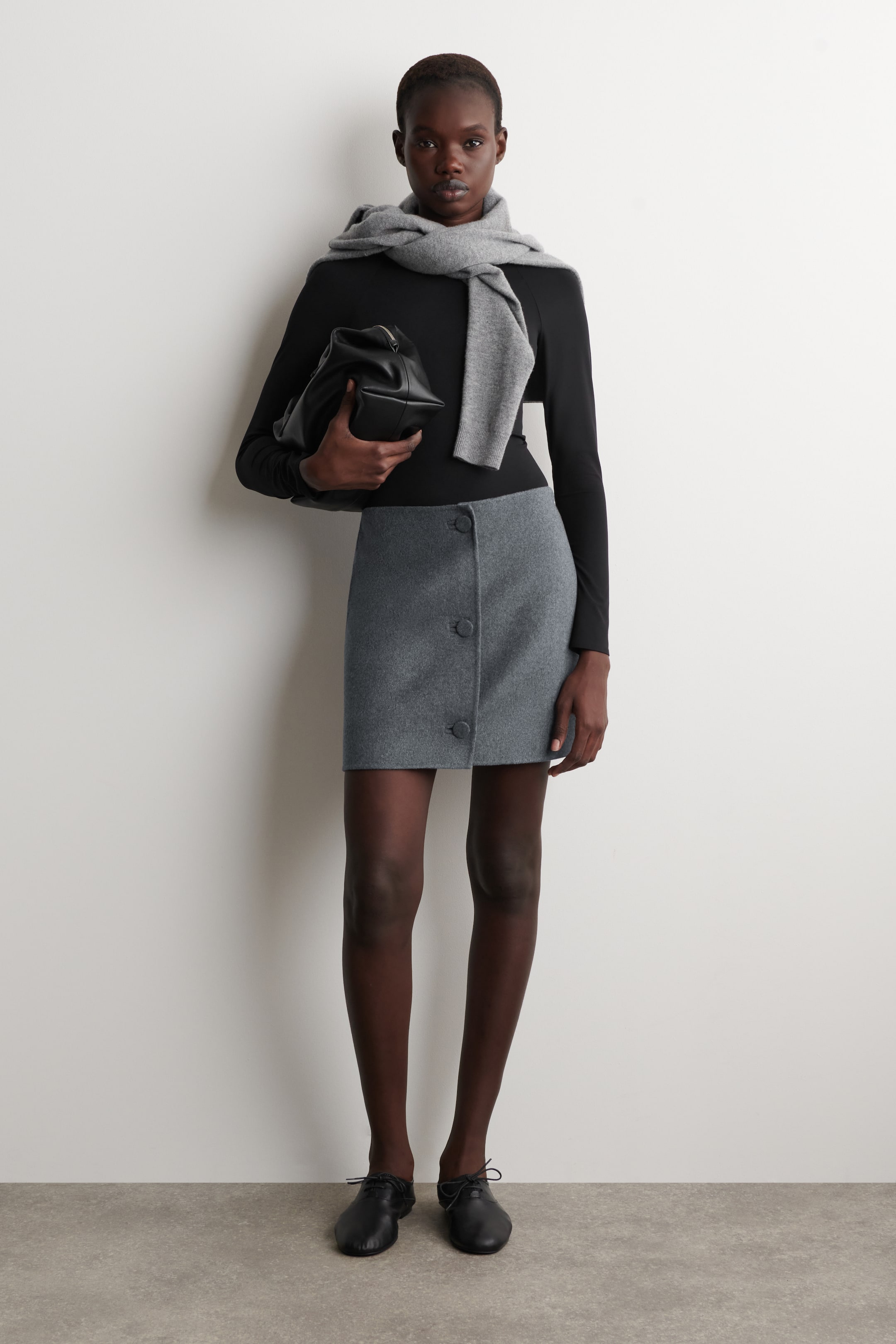 Vis større bilde: En grå miniskjørt med et subtilt teksturert stoff, en rett, over-kneet snitt, og tre runde, mørkegrå knapper vertikalt plassert på høyre side foran.