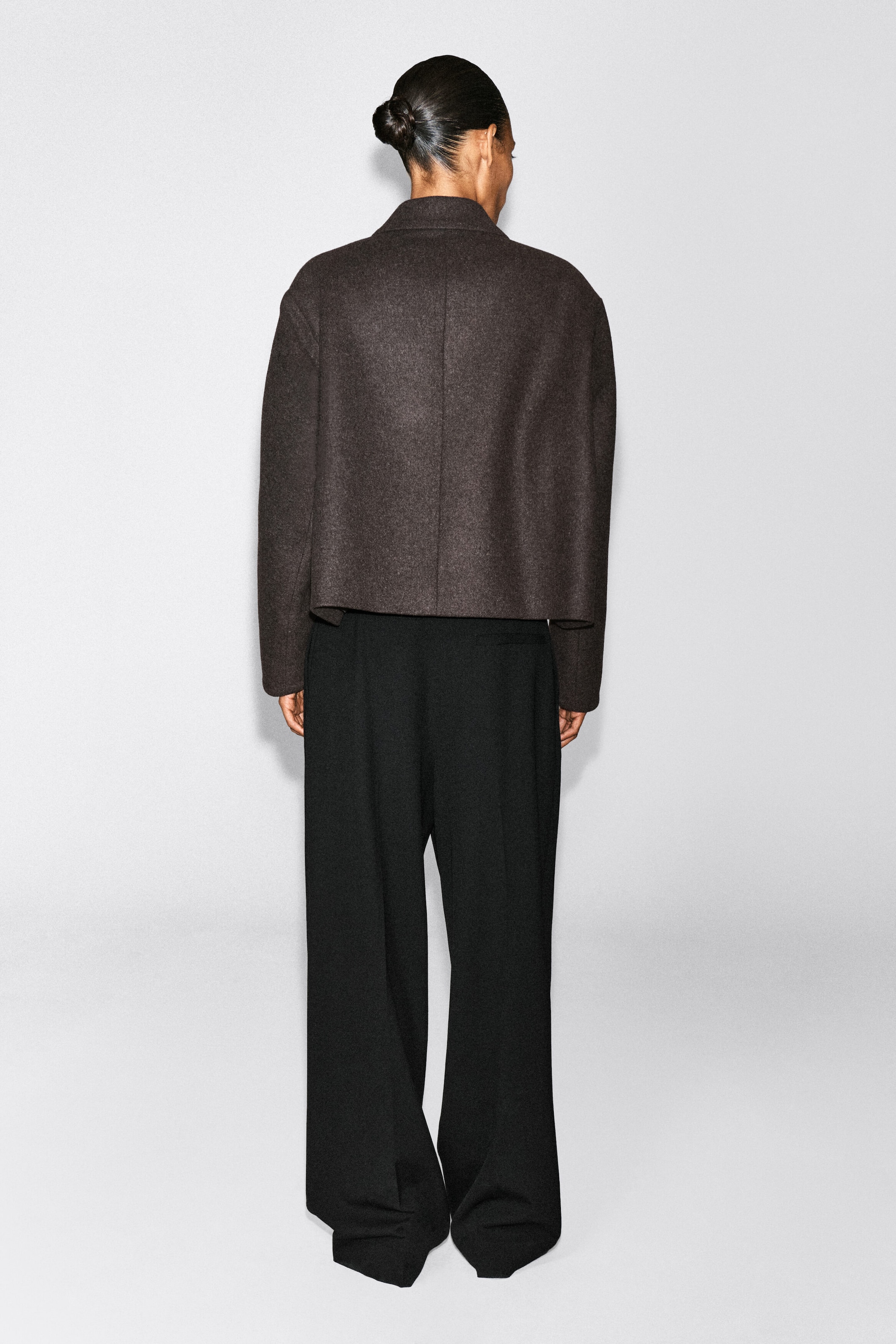 An oversized cropped boiled-wool blend blazer in dark brown is being worn by the model, viewed from behind. The boxy-fit blazer features a centre back seam and is paired with black wide-leg trousers. An oversized cropped boiled-wool blend blazer in dark brown is being worn by the model, viewed from behind. The boxy-fit blazer features a centre back seam and is paired with black wide-leg trousers.