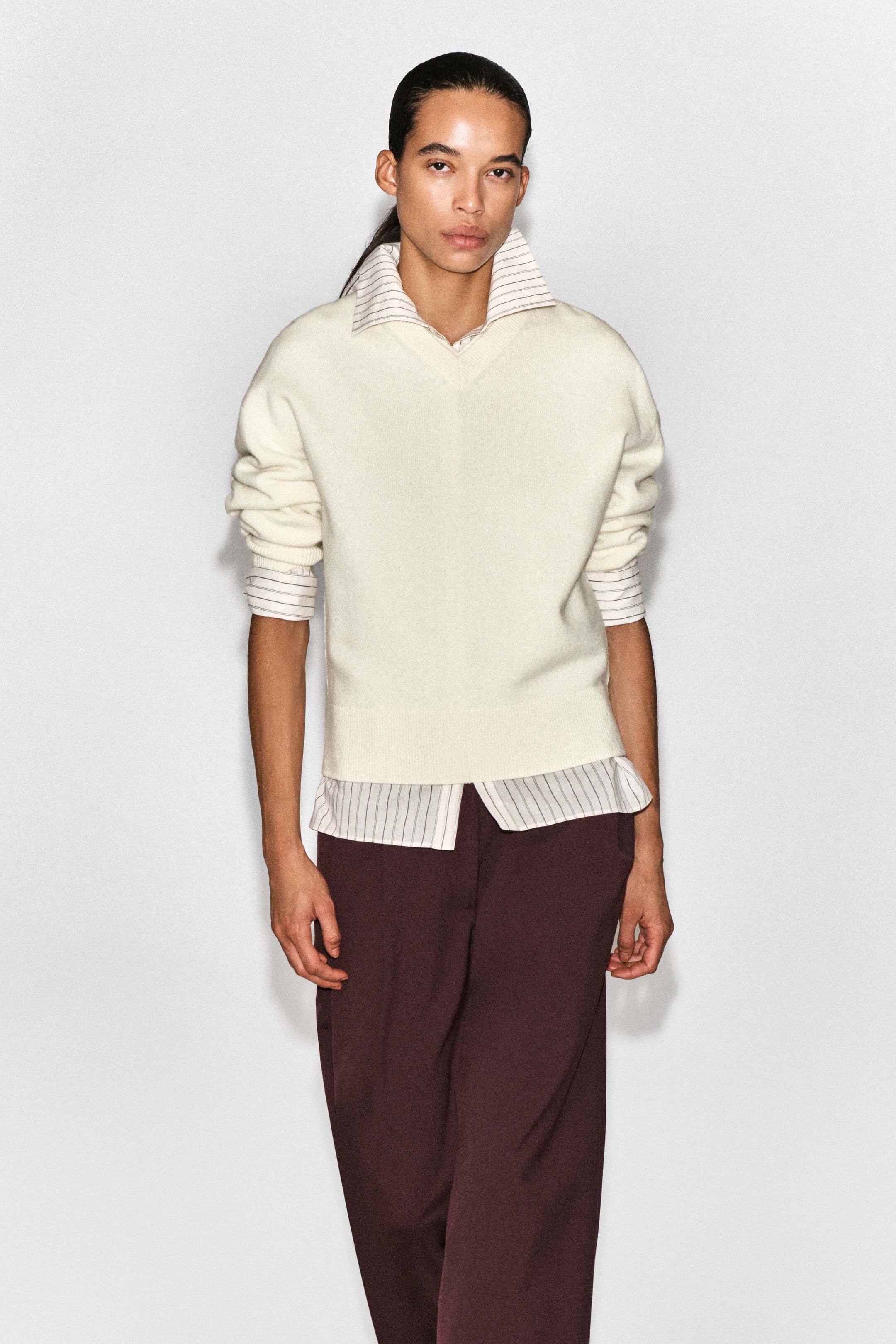 Agrandir l'image: Un mannequin porte le pull col V en laine mérinos couleur crème, présenté de face, avec une coupe décontractée, un col V, des poignets côtelés et un ourlet côtelé. Le pull est conçu avec une couche chemise rayée intégrée visible au col, aux poignets et à l’ourlet, créant un effet pré-stylé, associé à un pantalon bordeaux foncé.