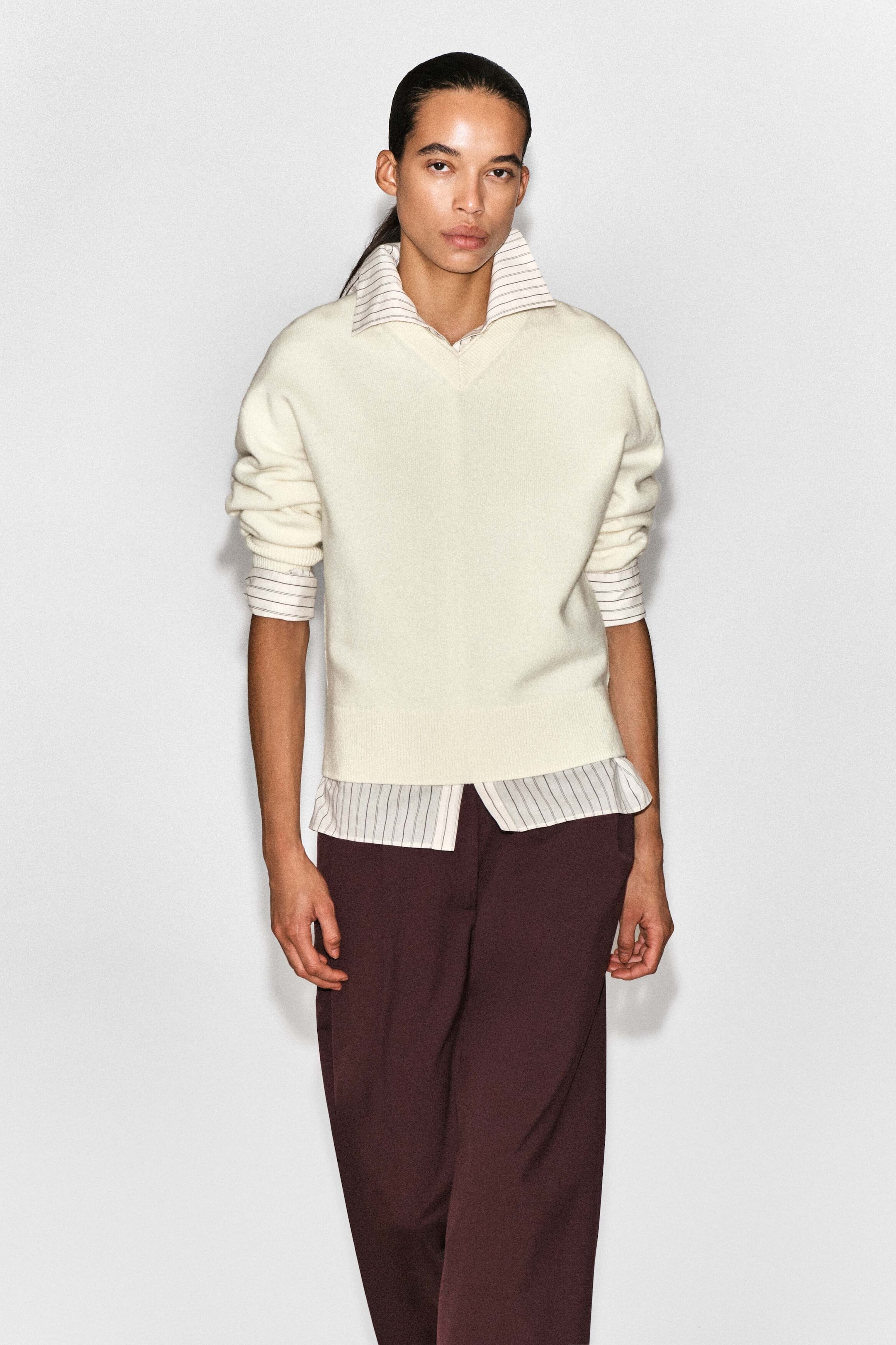 A model is wearing the cream-coloured Merino wool layered V-neck jumper, shown from the front, featuring a relaxed fit, a V-neckline, ribbed cuffs, and a ribbed hem. The jumper is designed with an integrated striped shirt layer visible at the collar, cuffs, and hem, creating a pre-styled look paired with dark burgundy trousers. A model is wearing the cream-coloured Merino wool layered V-neck jumper, shown from the front, featuring a relaxed fit, a V-neckline, ribbed cuffs, and a ribbed hem. The jumper is designed with an integrated striped shirt layer visible at the collar, cuffs, and hem, creating a pre-styled look paired with dark burgundy trousers.