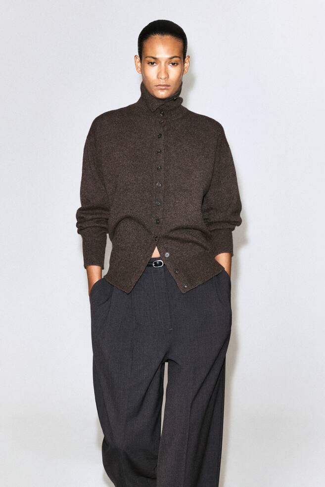 A dark brown knitted cardigan featuring a high funnel neck, front button fastening and ribbed cuffs is worn by the model. The cardigan is styled partially unbuttoned at the hem and paired with dark grey trousers. - 1