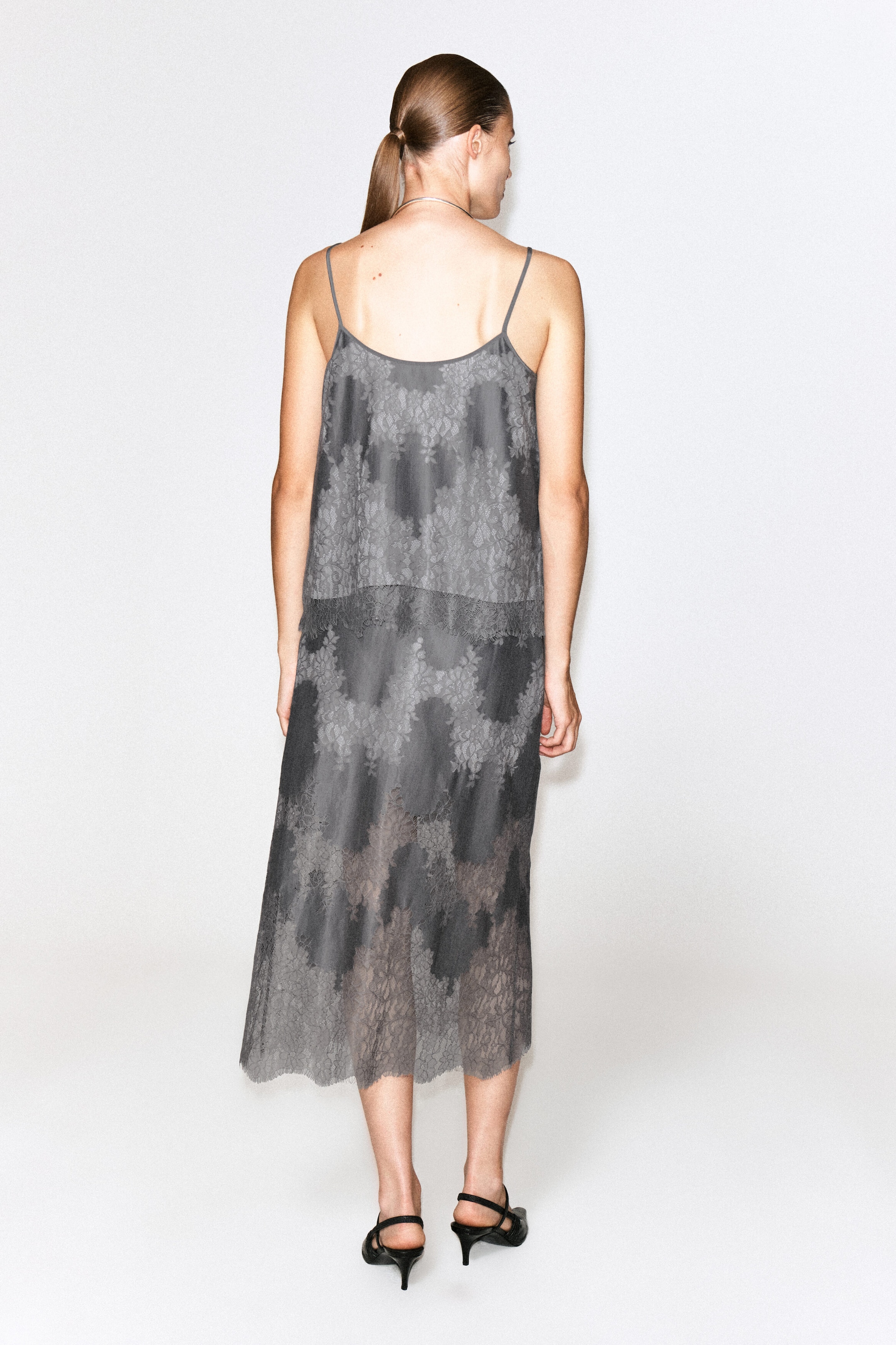 A grey lace-overlay cami top featuring thin spaghetti straps and a low scoop back is being worn by the model. The top is styled with a matching sheer lace midi skirt, creating a coordinated, layered look. A grey lace-overlay cami top featuring thin spaghetti straps and a low scoop back is being worn by the model. The top is styled with a matching sheer lace midi skirt, creating a coordinated, layered look.