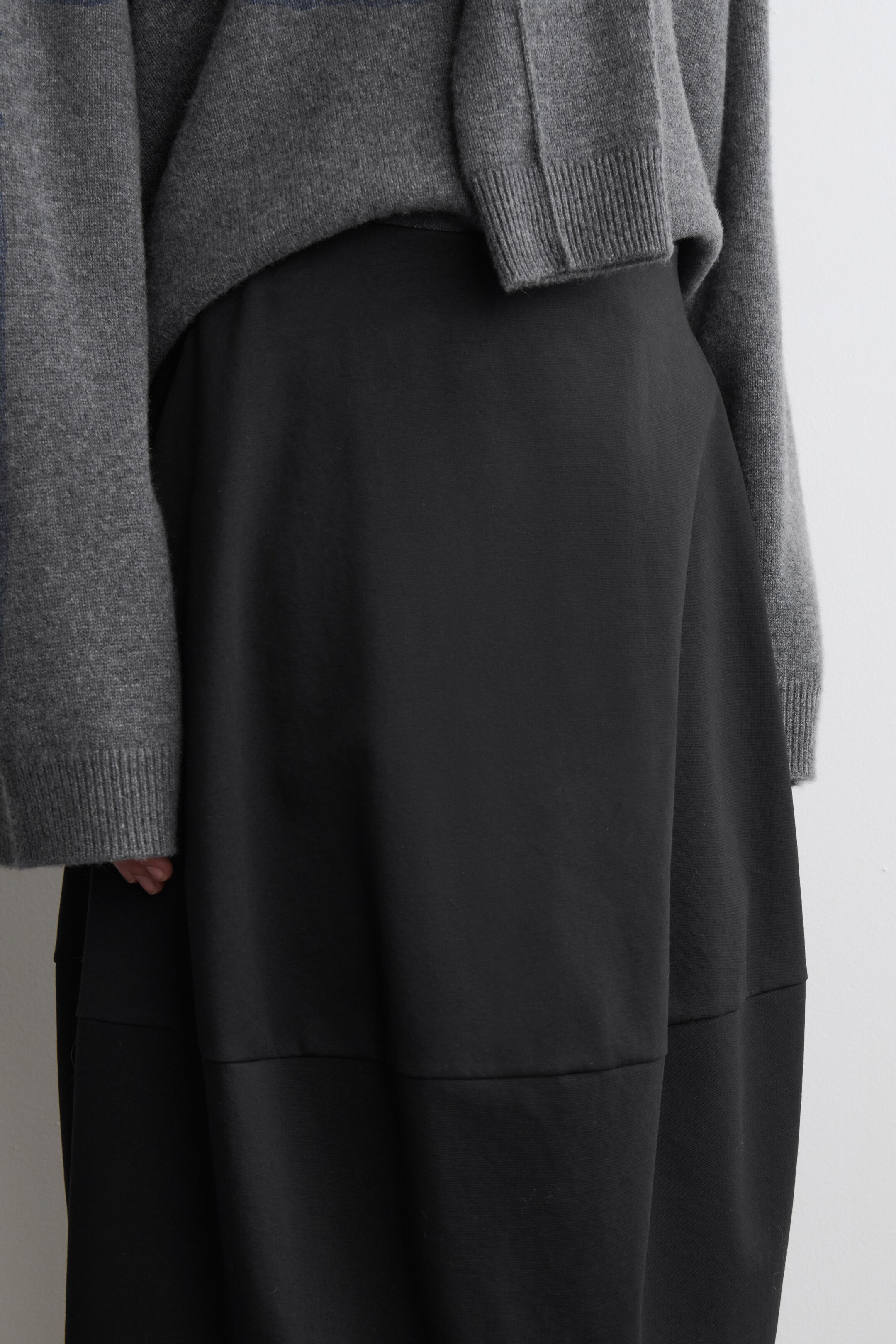 Agrandir l'image: Une jupe noire et fluide présente une texture lisse et mate ainsi qu'une large couture horizontale près de l'ourlet, indiquant un design à panneaux. Elle est portée en superposition sous un pull en tricot gris, qui présente des poignets côtelés et une coupe ample et décontractée.