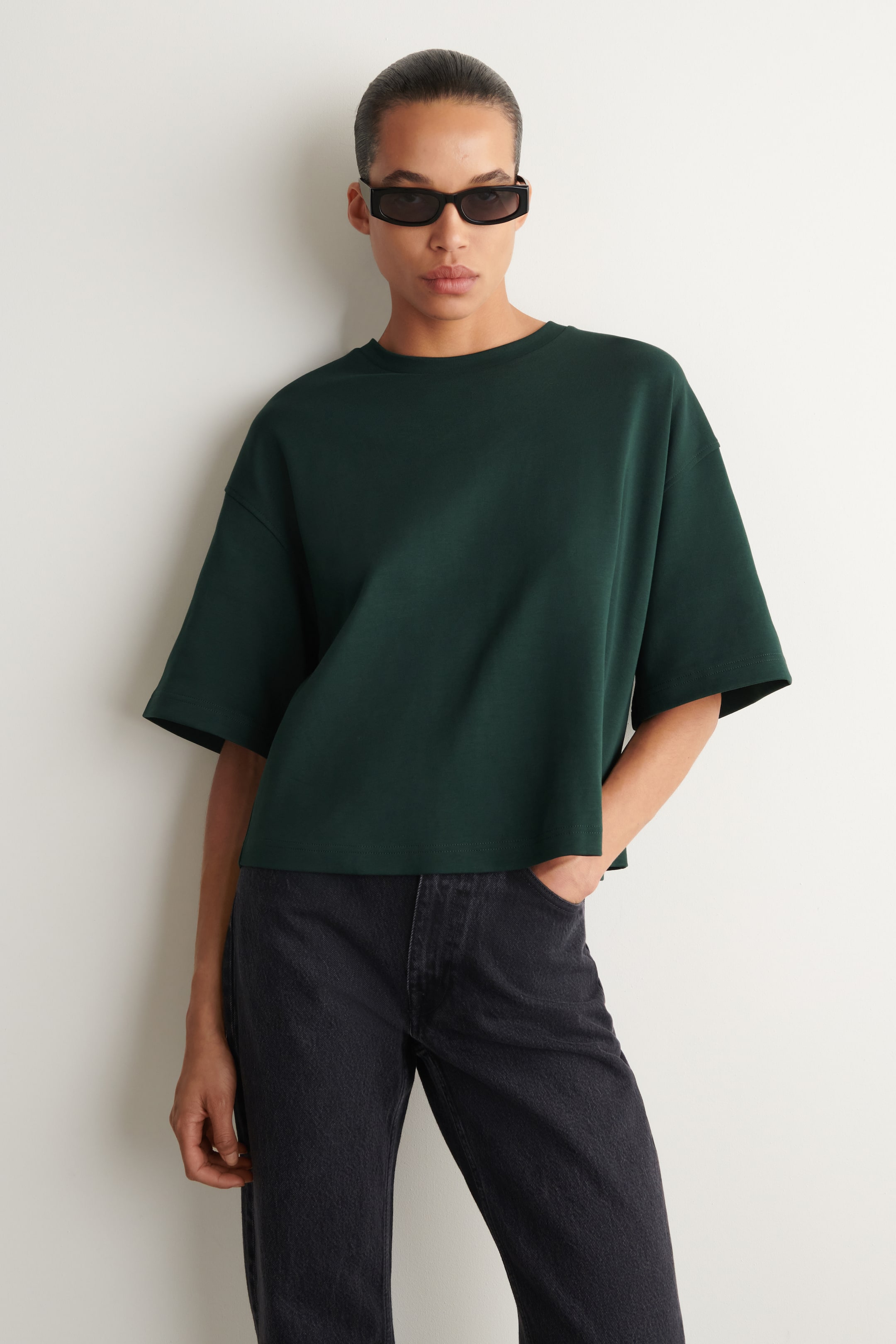 Ver imagem maior: Uma t-shirt oversized verde escura é apresentada de frente, destacando o seu corte amplo. Apresenta uma gola redonda canelada, mangas curtas caídas e uma bainha reta, ligeiramente cropped.