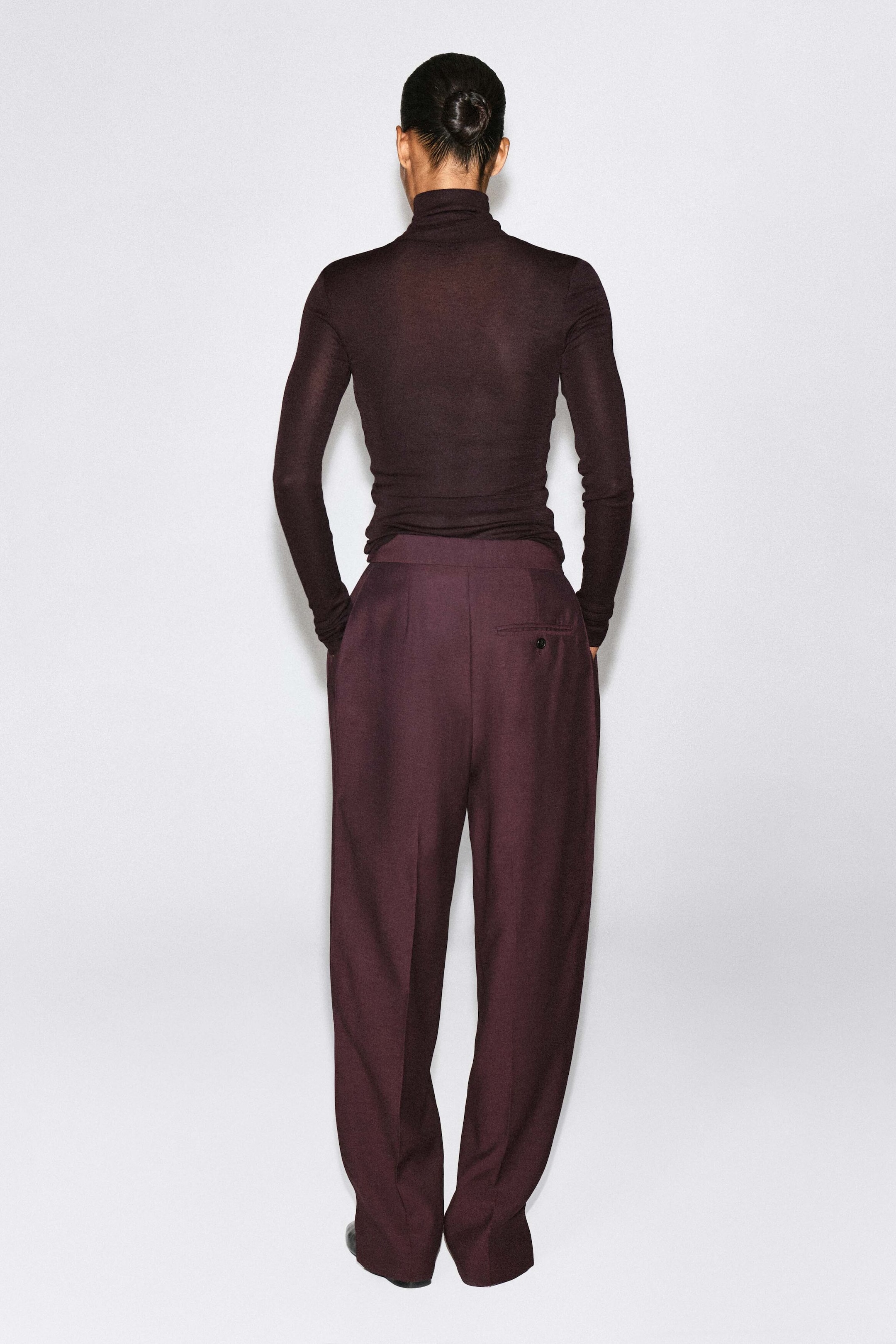 Pleated-panel straight-leg pants in a deep burgundy color are worn by the model, who is facing away from the camera. The pants feature pressed creases down the center of the legs and a single buttoned pocket on the back, paired with a tucked-in, dark long-sleeve turtleneck top. Pleated-panel straight-leg pants in a deep burgundy color are worn by the model, who is facing away from the camera. The pants feature pressed creases down the center of the legs and a single buttoned pocket on the back, paired with a tucked-in, dark long-sleeve turtleneck top.