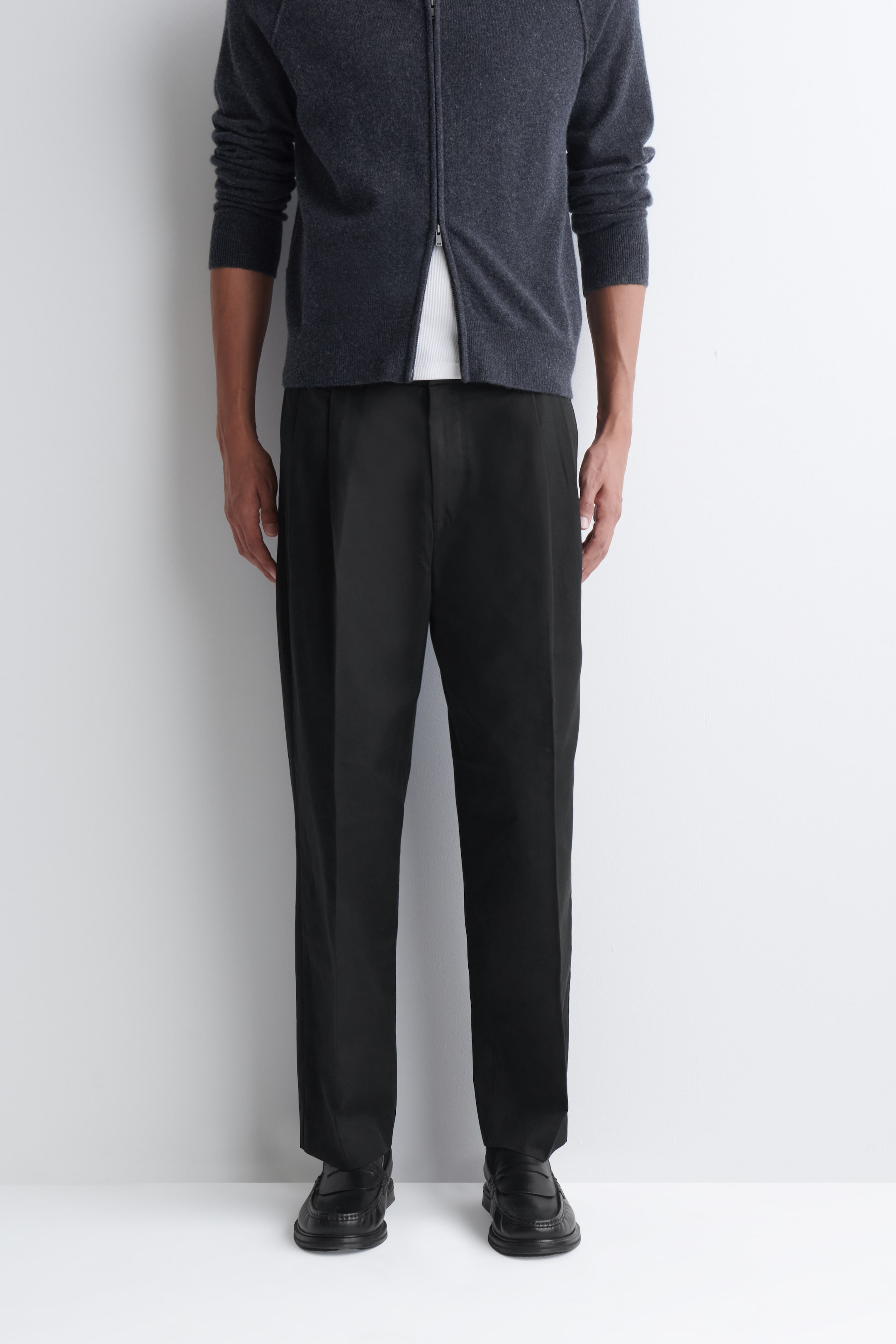 Visualizza immagine più grande: Pantaloni neri a gamba affusolata con vestibilità relaxed e pieghe sono indossati dalla modella. I pantaloni presentano pieghe visibili in vita, una vestibilità rilassata lungo la gamba e un fondo gamba affusolato alla caviglia, e sono abbinati a un cardigan grigio scuro e a mocassini neri.