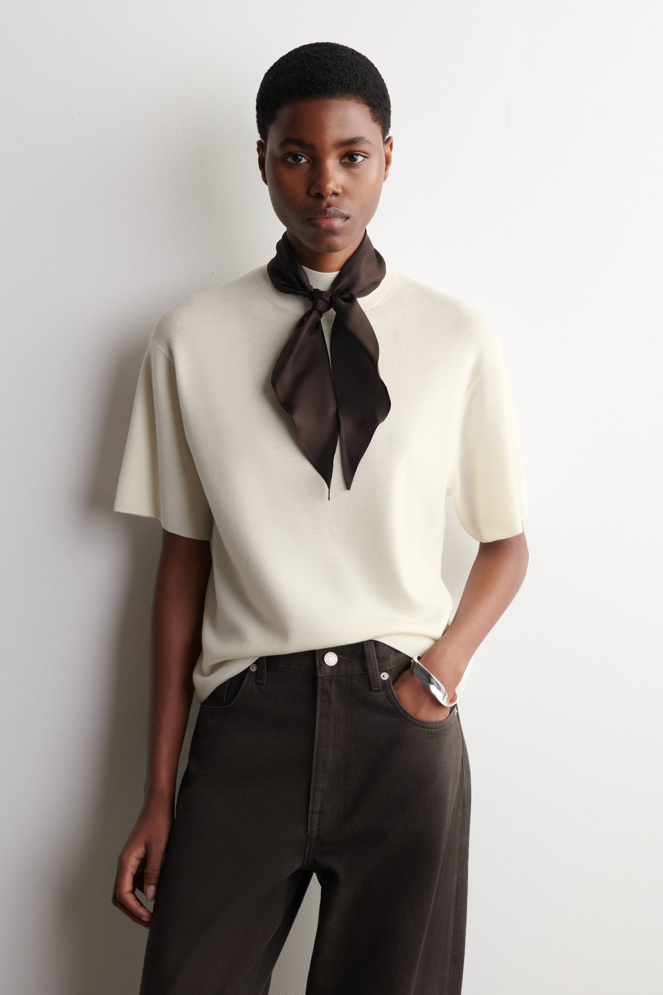 Agrandir l'image: Un haut en maille à manches courtes crème est présenté, mettant en valeur sa coupe décontractée et son col rond haut. Un foulard en satin marron foncé est noué au cou, avec ses extrémités tombant sur le haut. Un pantalon marron foncé à taille haute avec des poches avant visibles est porté, affichant sa coupe droite.