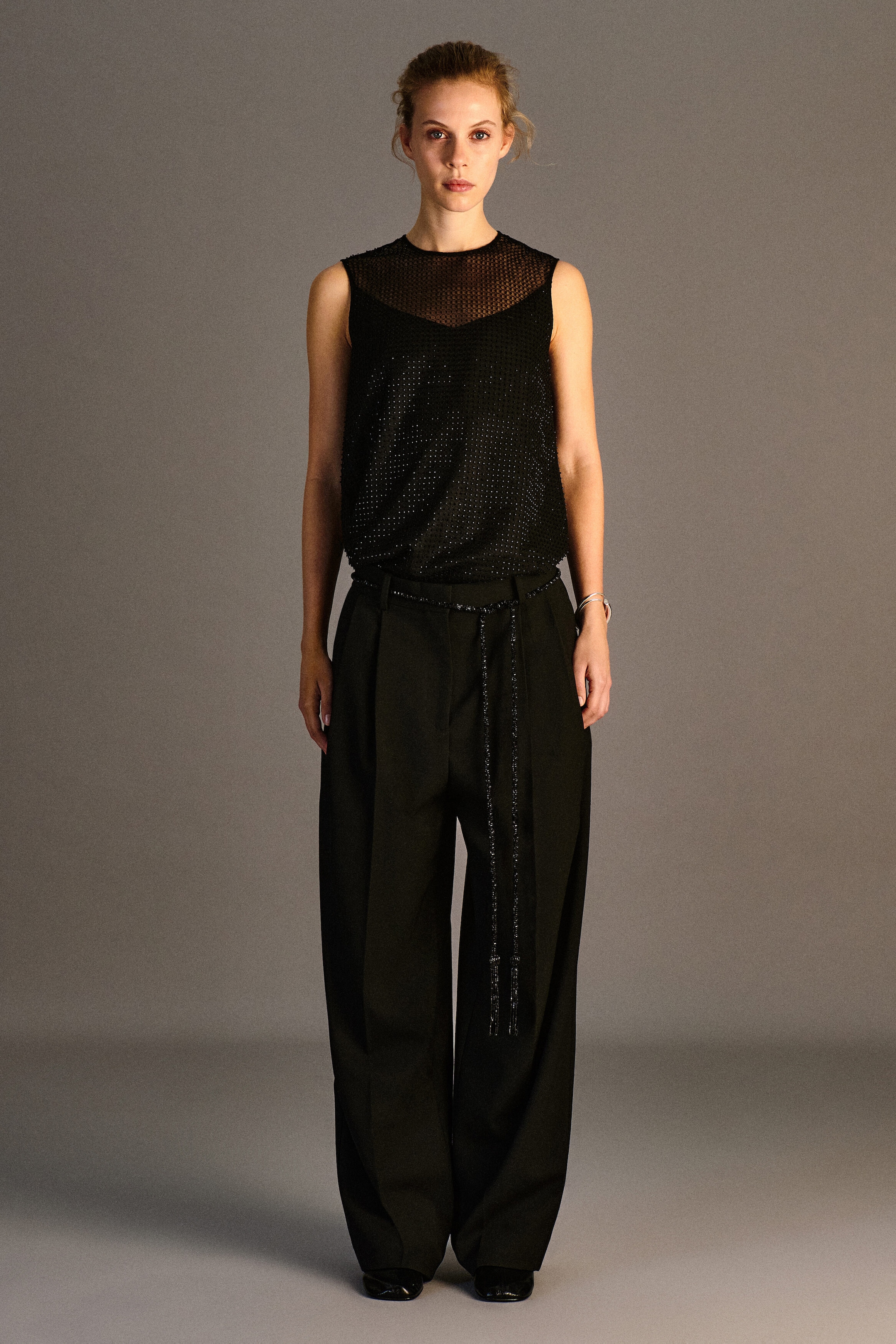 Ein schwarzes, mit Perlen verziertes Stricktop ist frontal zu sehen und zeigt ein ärmelloses Design mit einem transparenten Mesh-Einsatz am Schulterbereich und Rundhalsausschnitt. Der Körper des Oberteils ist mit dezenten, schimmernden Perlenverzierungen versehen und in eine schwarze Hose gesteckt, um die lockere Passform zu betonen. Ein schwarzes, mit Perlen verziertes Stricktop ist frontal zu sehen und zeigt ein ärmelloses Design mit einem transparenten Mesh-Einsatz am Schulterbereich und Rundhalsausschnitt. Der Körper des Oberteils ist mit dezenten, schimmernden Perlenverzierungen versehen und in eine schwarze Hose gesteckt, um die lockere Passform zu betonen.