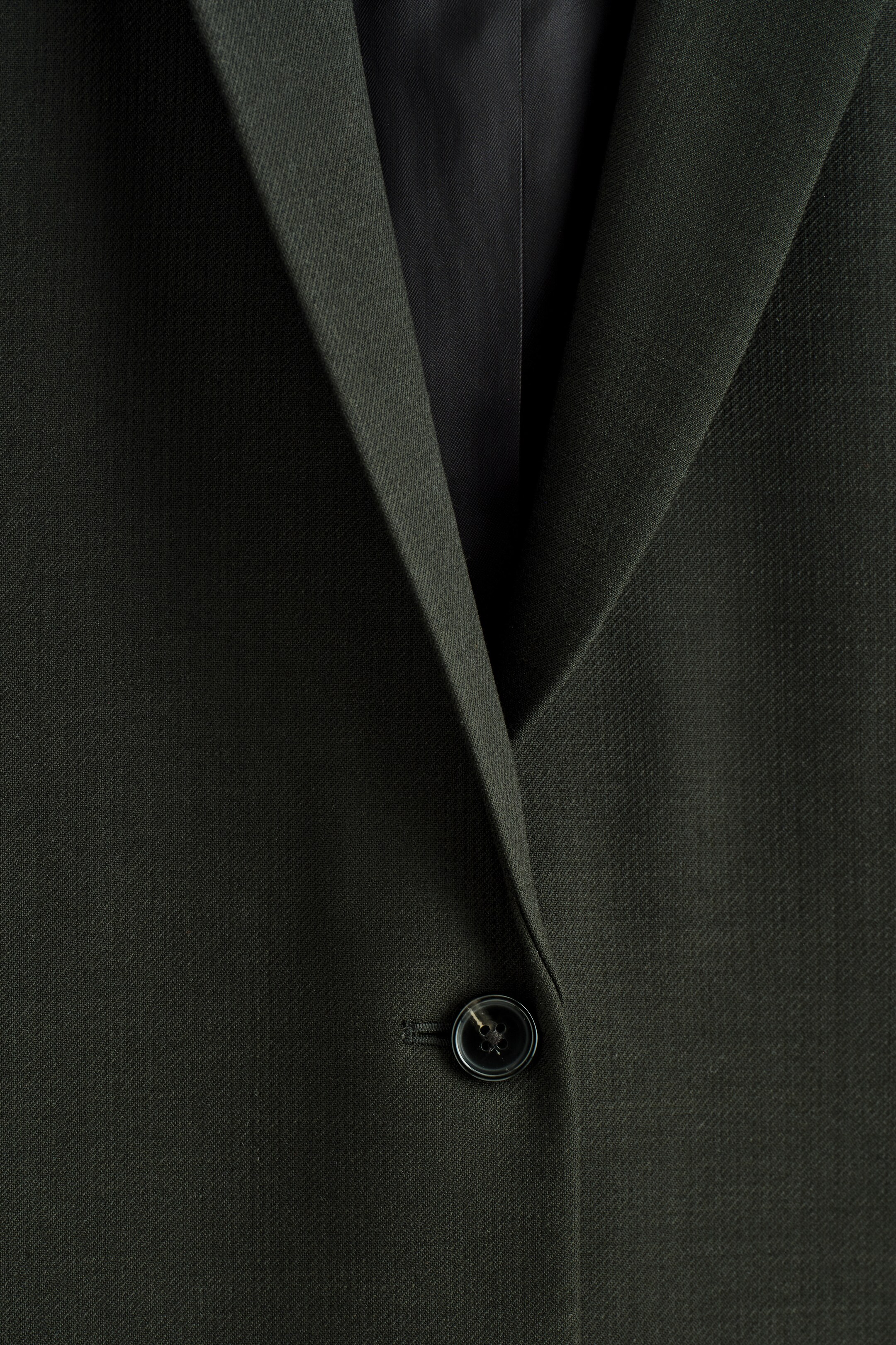 Grotere afbeelding bekijken: Een diep-groene blazer wordt van voren getoond, met een single-breasted sluiting en een zichtbare donkere knop. De blazer is gemaakt van een subtiel getextureerde stof en de notch revers vormen een V-hals opening.