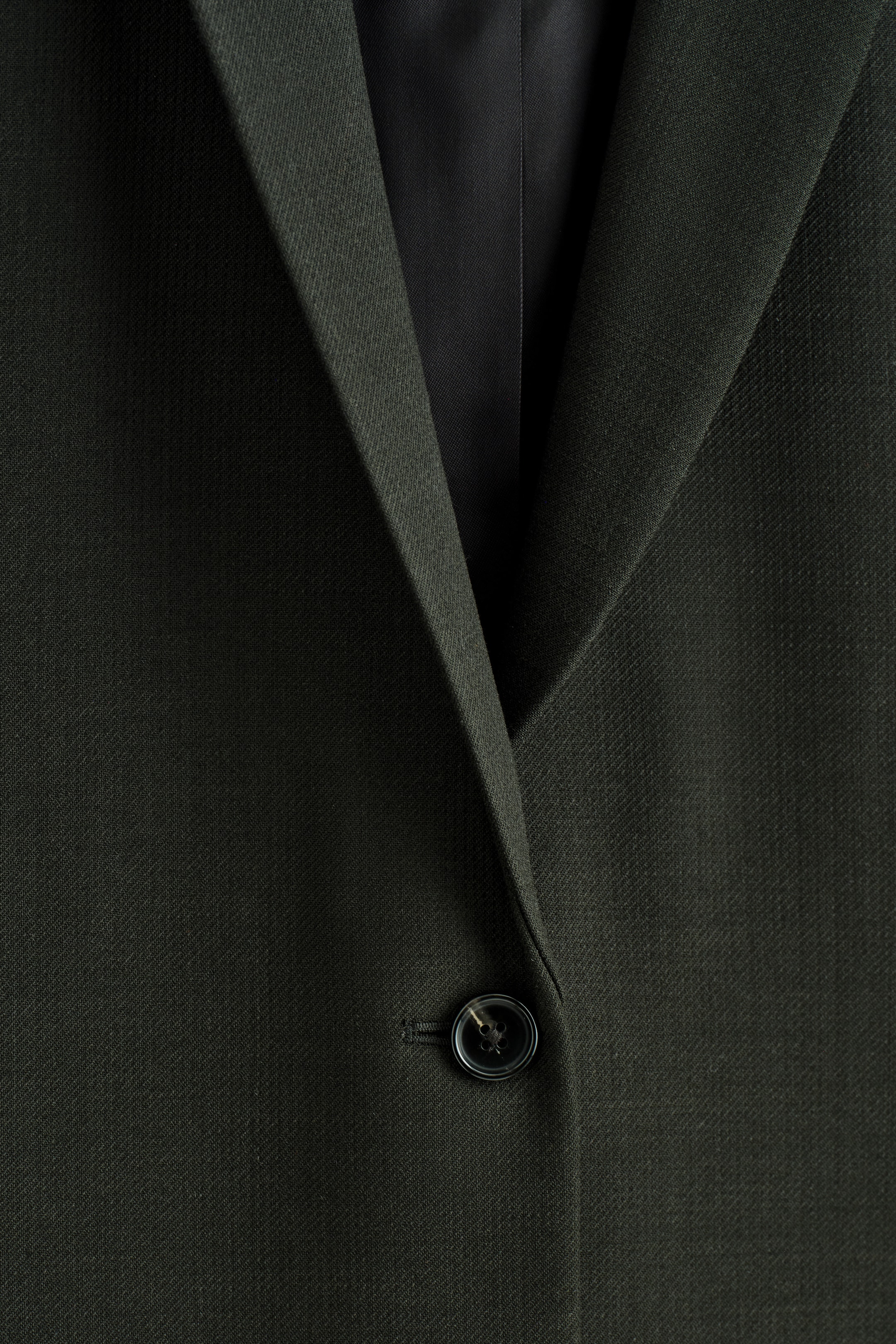 Grösseres Bild ansehen: Ein tiefgrüner Blazer wird von vorne gezeigt, mit einer einreihigen Schließung und einem sichtbaren dunklen Knopf. Der Blazer ist aus einem dezent strukturierten Stoff gefertigt, und seine steigenden Revers formen einen V-Ausschnitt.