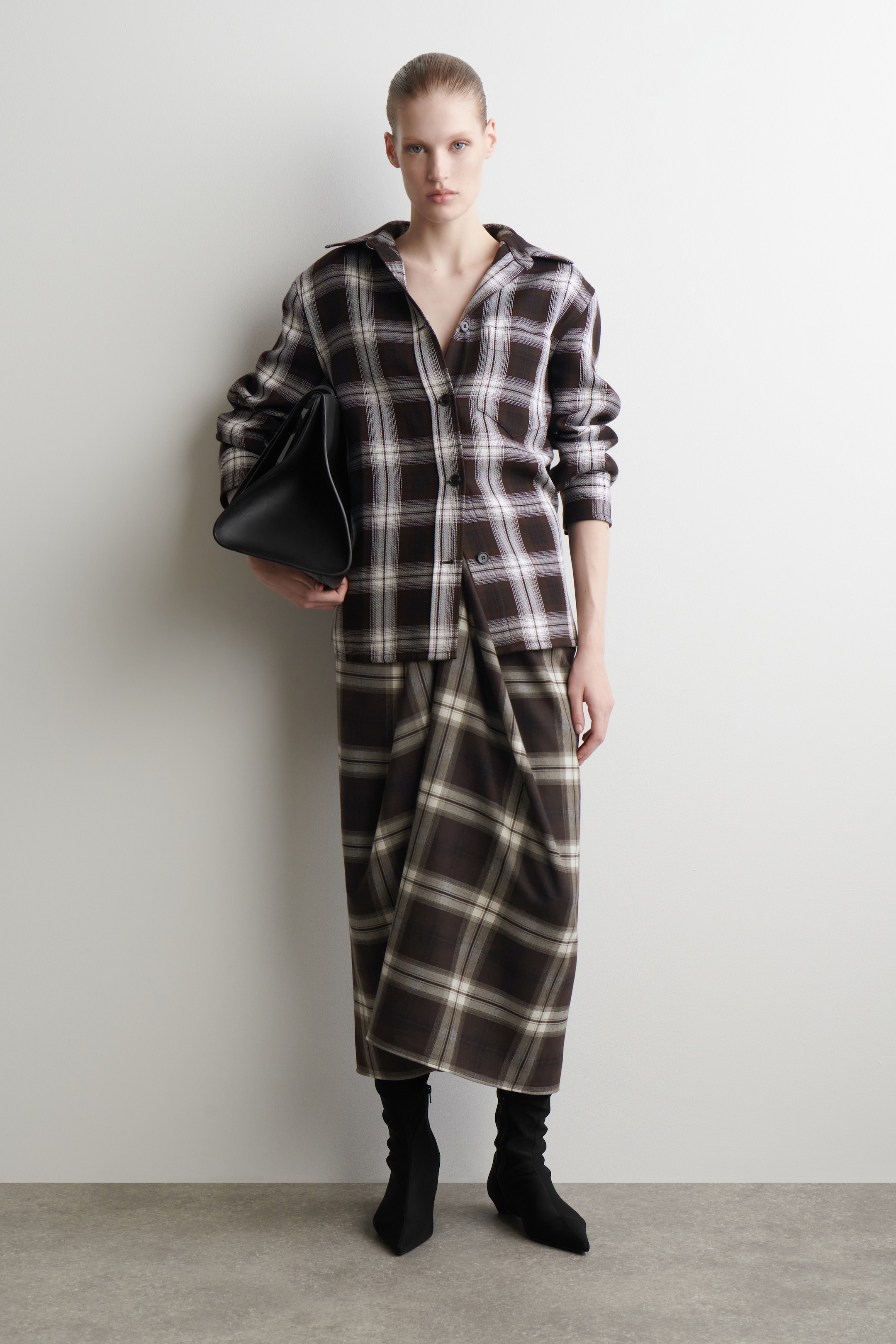 Agrandir l'image: Un ensemble de chemise et jupe à carreaux marron, blanc et noir est présenté, face vers l'avant. La chemise oversize à boutons présente un col ouvert et des manches longues, tandis que la jupe longue affiche un détail drapé à l'avant. Un sac à main structuré noir est tenu, et des bottines noires à bout pointu complètent le look.