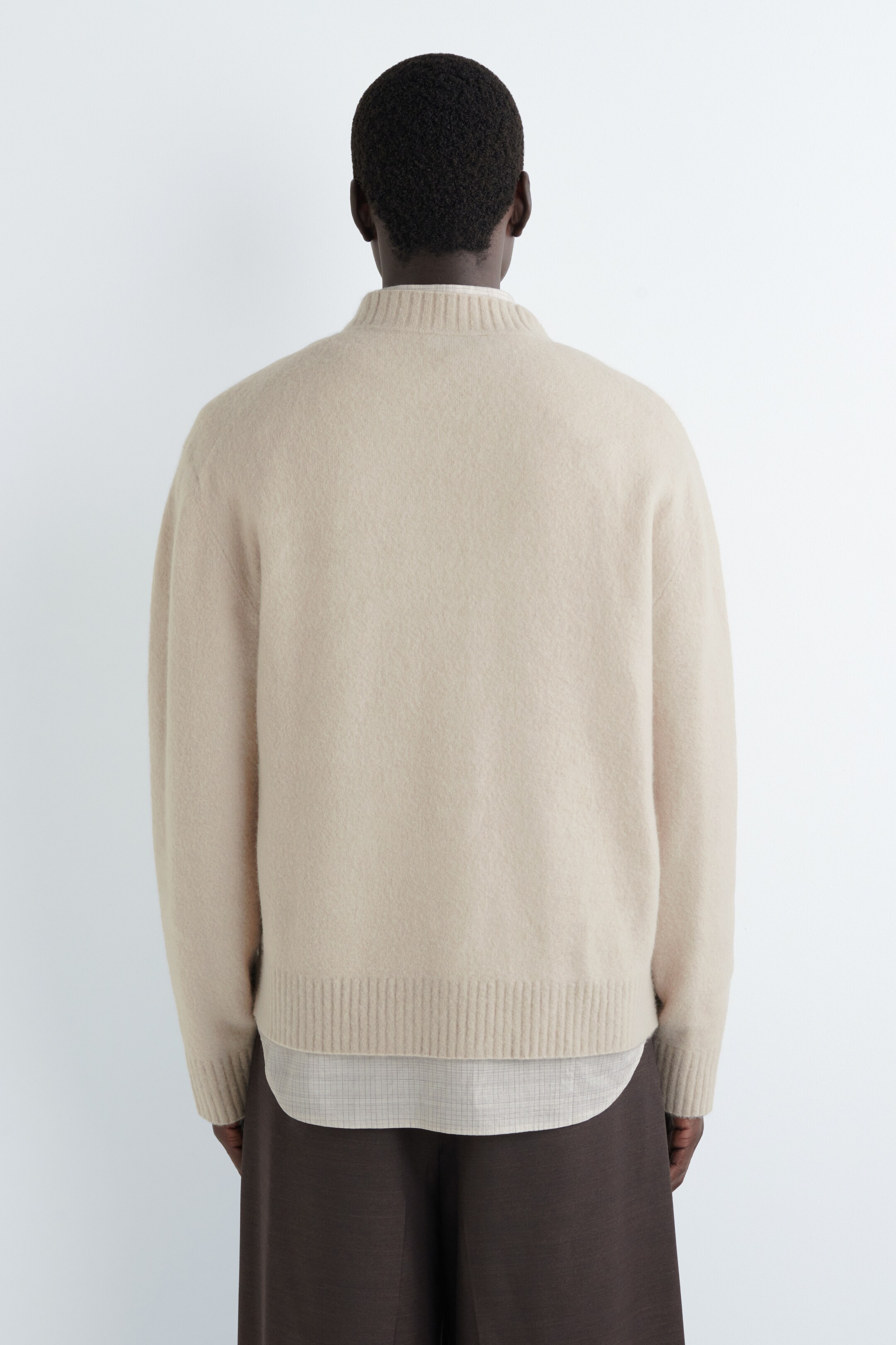 Ver imagen más grande: Un jersey beige se está viendo desde atrás, con un cuello alto acanalado, mangas largas con puños acanalados y un dobladillo acanalado. El suave jersey de punto se está superponiendo sobre una camisa ligera con un sutil patrón, que se extiende más allá de su dobladillo.