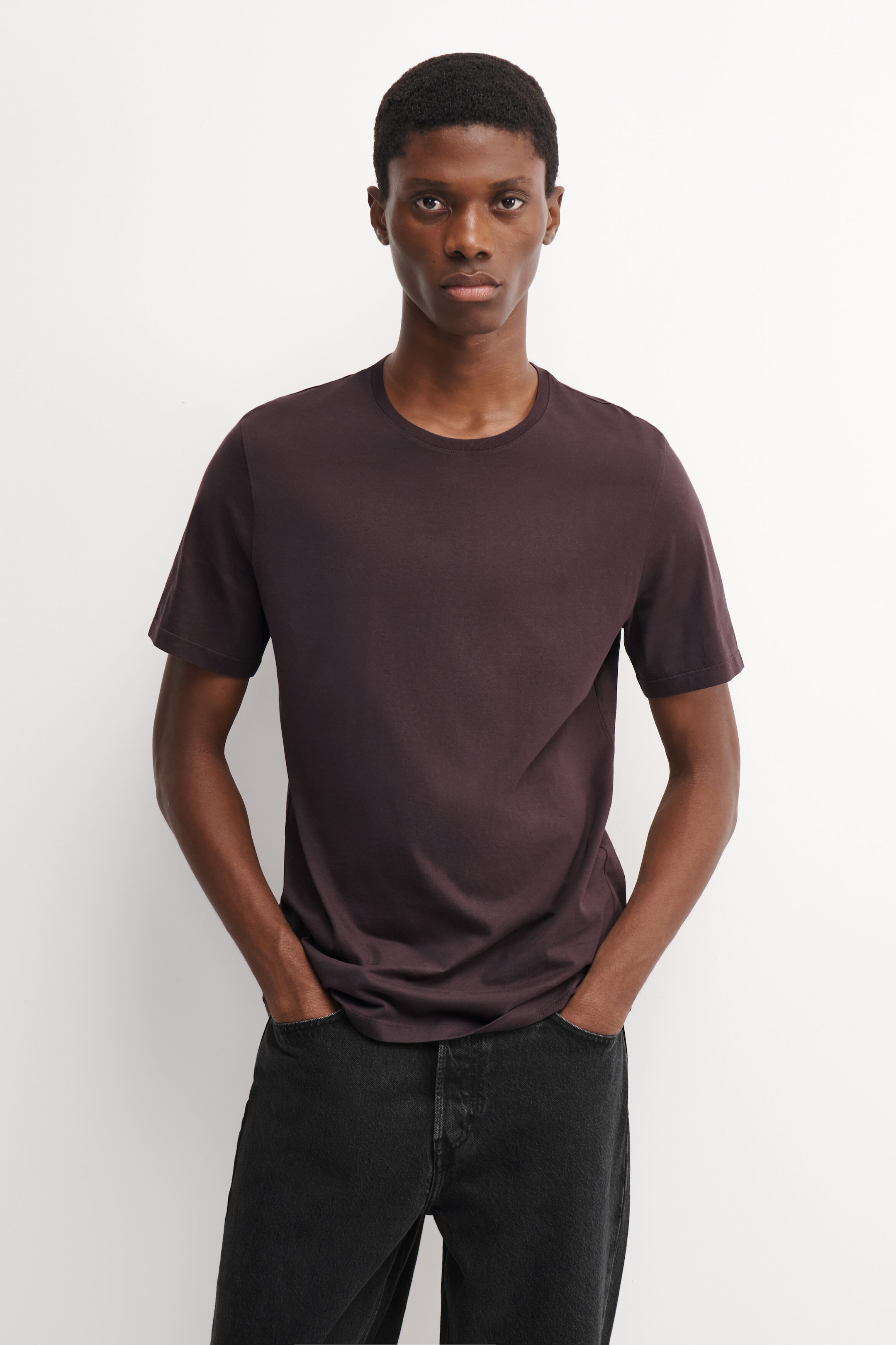 The model is wearing a dark plum cotton crew-neck T-shirt, paired with dark jeans and standing with her hands in her pockets. The T-shirt features a classic fit, short sleeves, and a clean crew neckline. The model is wearing a dark plum cotton crew-neck T-shirt, paired with dark jeans and standing with her hands in her pockets. The T-shirt features a classic fit, short sleeves, and a clean crew neckline.