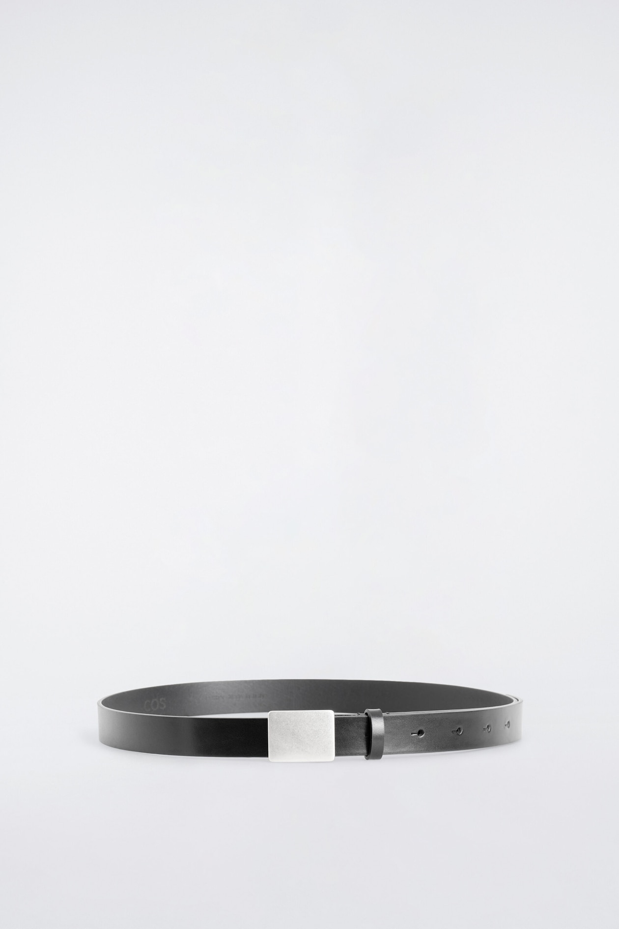 A black leather belt is resting in a circular loop, facing front to showcase its structure. The belt features a smooth black leather strap, a square brushed silver-toned buckle, and a matching leather keeper loop.