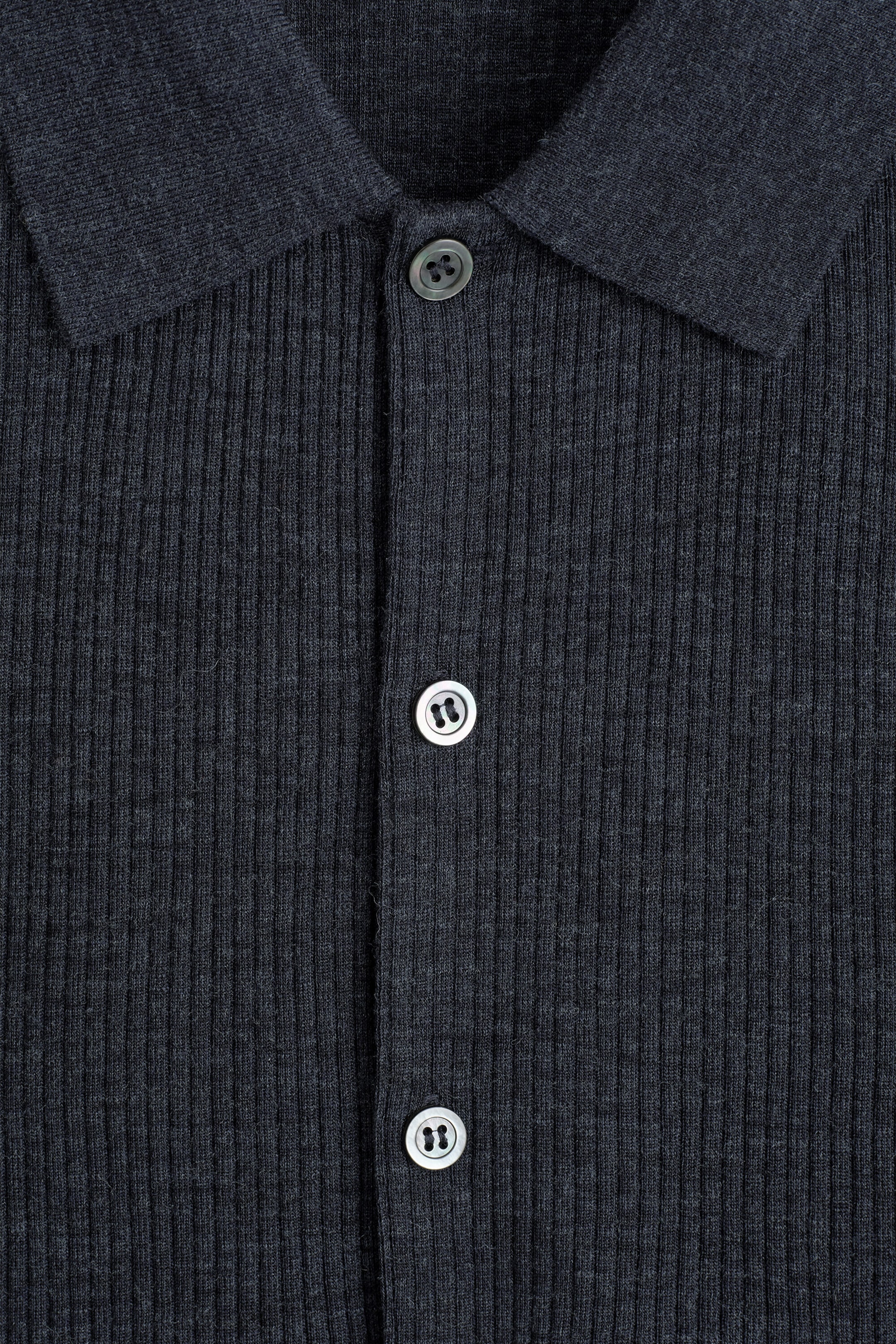 A close-up front view of a dark grey ribbed knit shirt shows a classic flat collar and a central button placket. The shirt features a dark grey top button and two contrasting light grey buttons aligned down the placket.