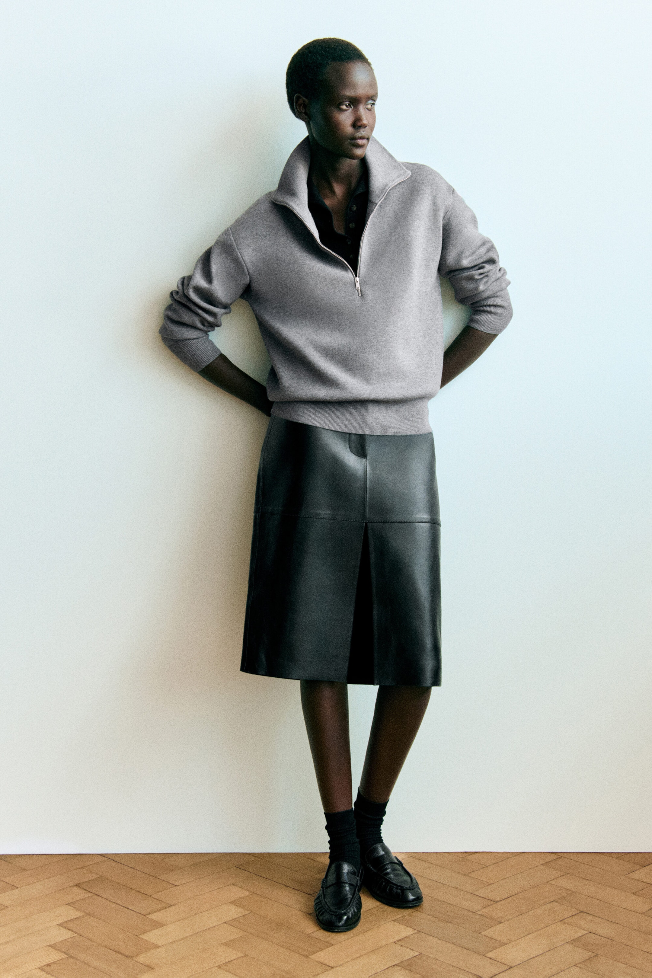 A model is wearing a gray funnel-neck half-zip knit sweater and a black leather midi skirt in a front-facing view. The sweater features ribbed trims and is layered over a dark collared shirt, while the structured leather skirt shows a front seam and a subtle center slit. A model is wearing a gray funnel-neck half-zip knit sweater and a black leather midi skirt in a front-facing view. The sweater features ribbed trims and is layered over a dark collared shirt, while the structured leather skirt shows a front seam and a subtle center slit.