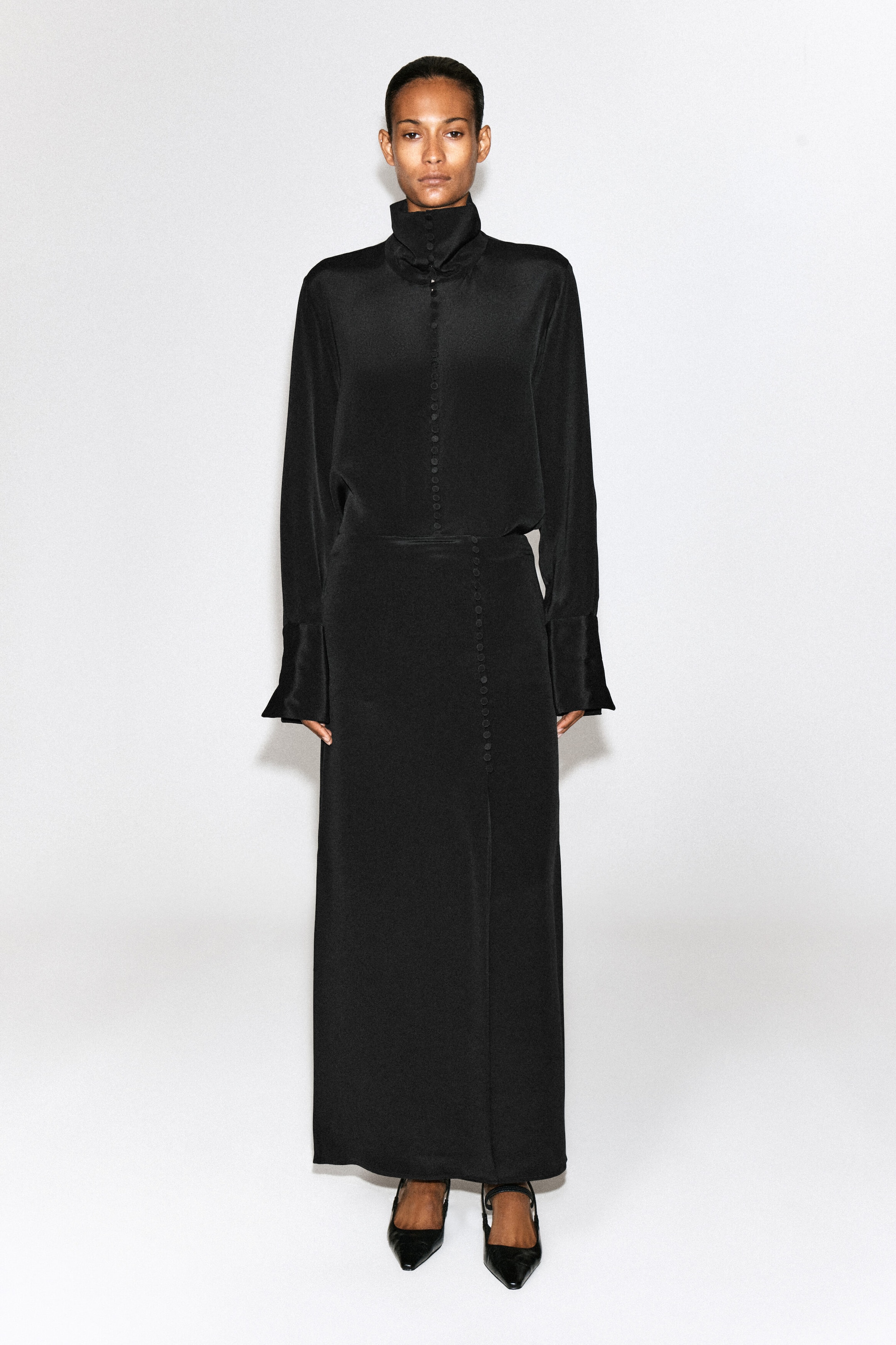 The model is wearing a black funnel-neck silk shirt featuring a high collar, a central row of small fabric-covered buttons, and long sleeves with wide cuffs. The shirt is styled tucked into a matching long black skirt. The model is wearing a black funnel-neck silk shirt featuring a high collar, a central row of small fabric-covered buttons, and long sleeves with wide cuffs. The shirt is styled tucked into a matching long black skirt.