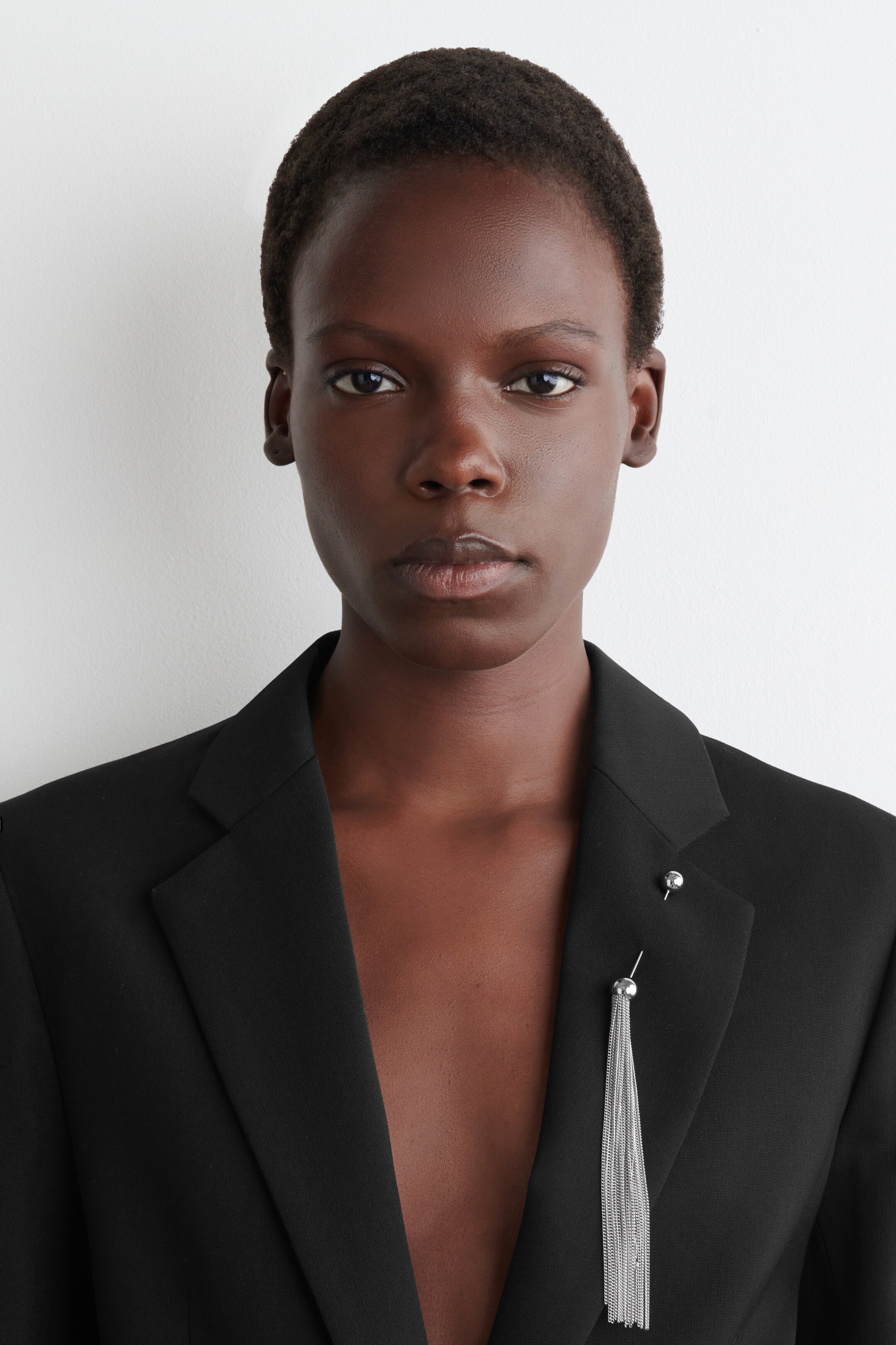 Ver imagen más grande: Un blazer negro presenta solapas con muescas en su lado derecho frontal. Dos alfileres decorativos de tono plateado adornan la solapa derecha, con un alfiler que presenta un diseño esférico y el otro que presenta una parte superior esférica con múltiples cadenas en cascada formando un fleco.
