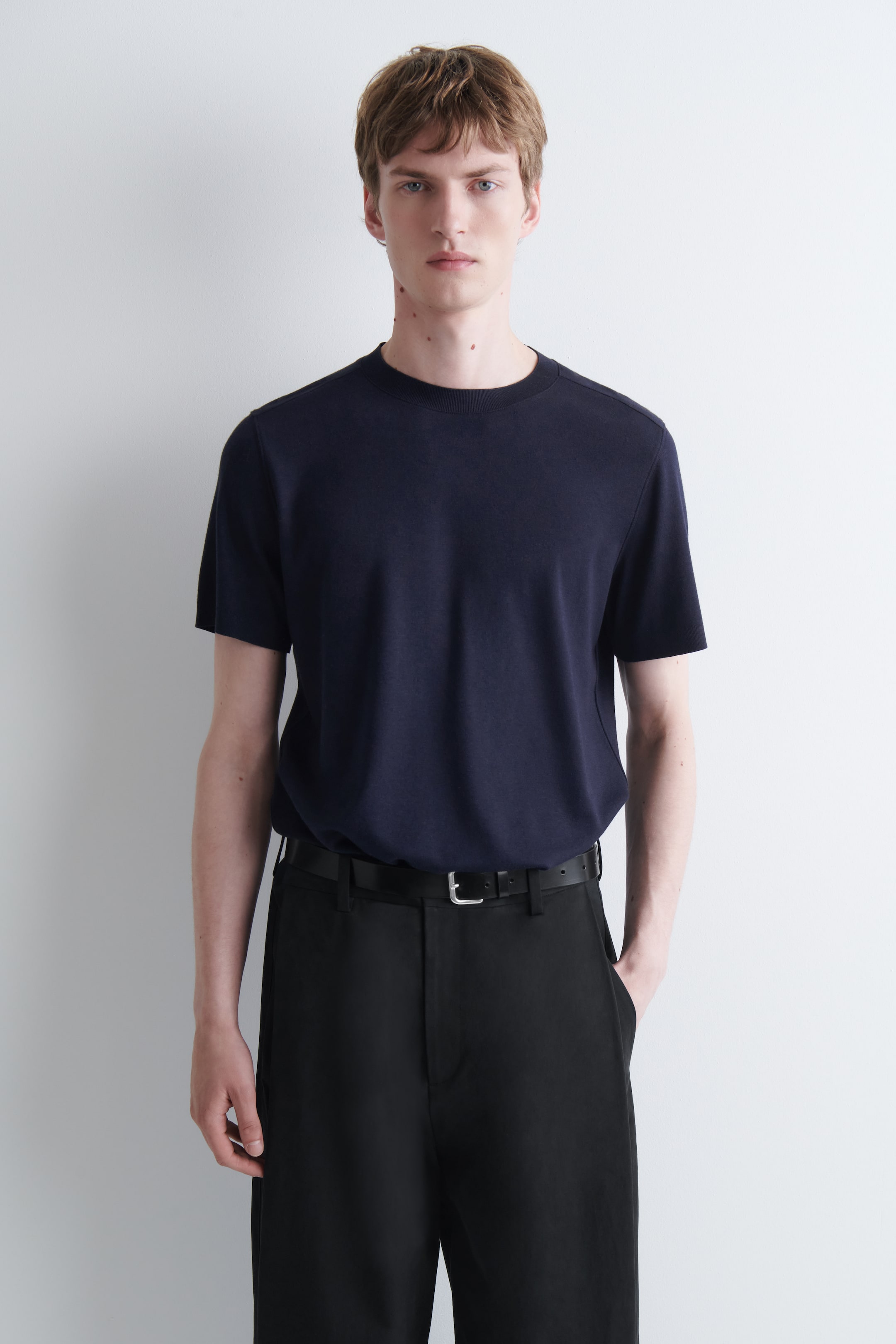 Visualizza l'immagine ingrandita: Una t-shirt lavorata a maglia di colore blu scuro con girocollo a coste e maniche corte è indossata dalla modella. La t-shirt è infilata in pantaloni neri, fissati con una cintura nera.