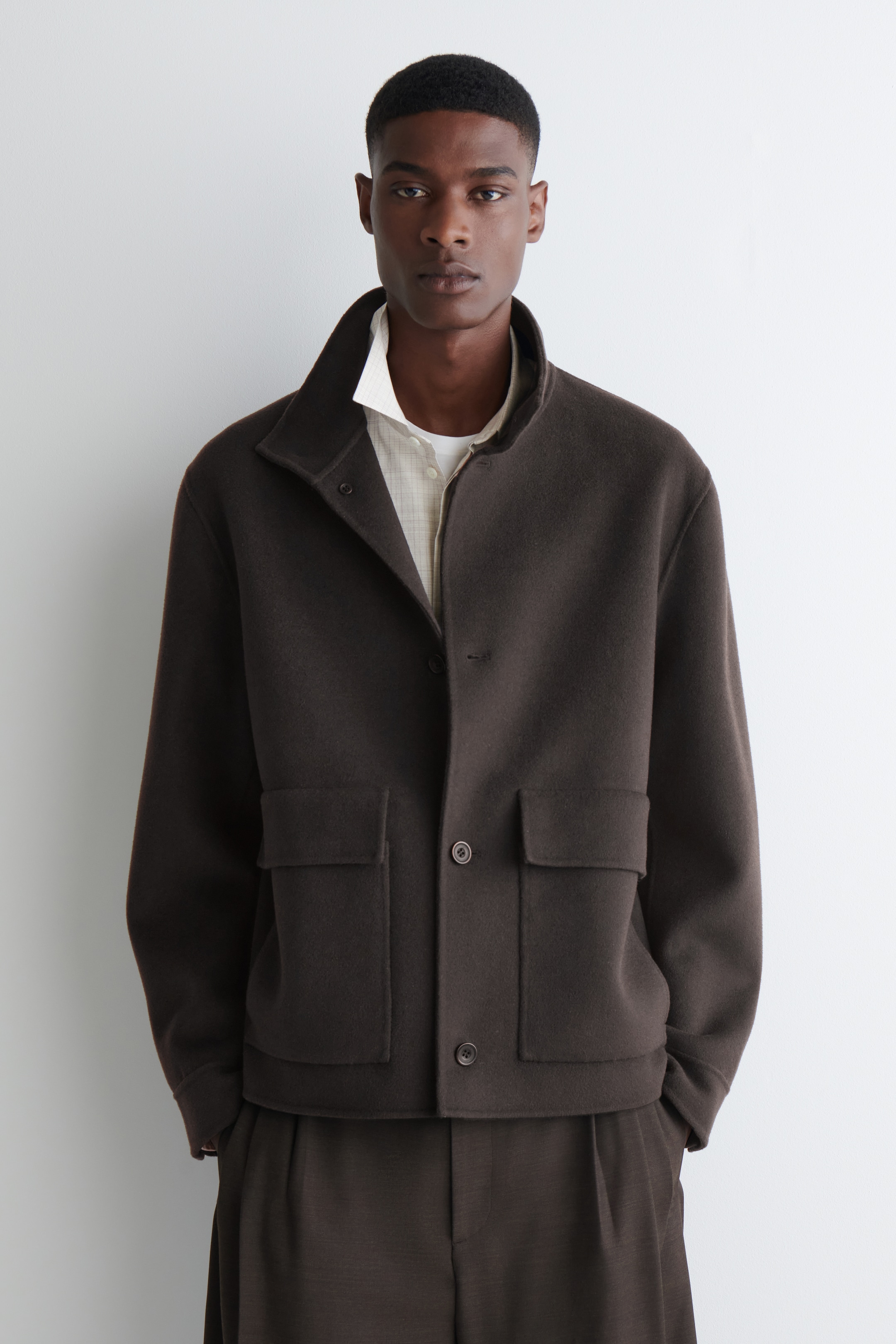 Grösseres Bild ansehen: Eine dunkelbraune Jacke mit einem Trichterkragen, einer Knopfleiste vorne und zwei großen Klappentaschen wird vom Model getragen. Die Hände des Models sind in den Taschen der Jacke, und darunter ist ein helles Hemd zu sehen.