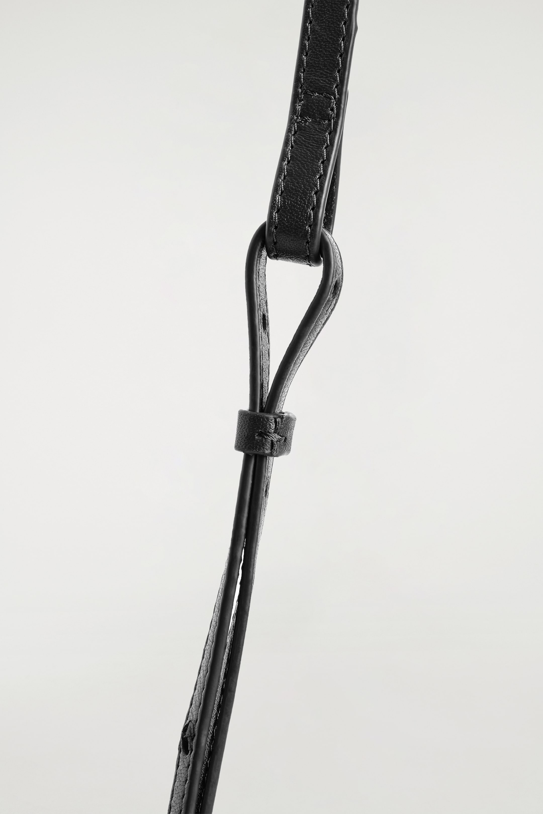 View larger image: The black leather strap of the TROVE CROSSBODY BAG - LEATHER is being shown in a close-up, detailed with visible edge stitching and a textured surface. The strap is looping through a dark ring and being secured by a small leather keeper below it.