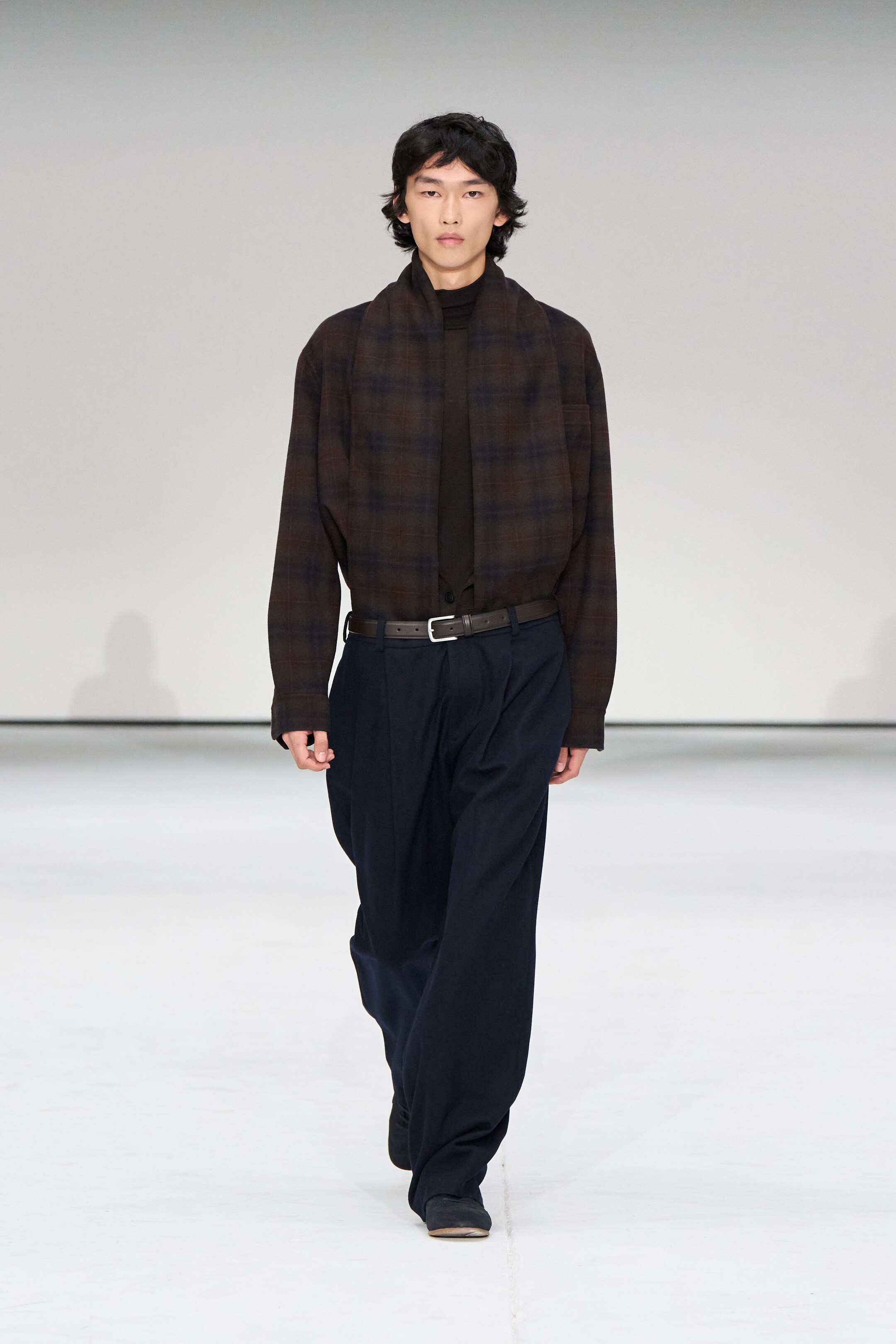 Größeres Bild anzeigen: Ein Overshirt mit entspannter Passform in einem dunkelbraunen Karomuster, das dunkelrote und dunkelblaue Linien aufweist, wird über einem dunklen Rollkragenpullover getragen und zeigt einen spitzen Kragen und geknöpfte Manschetten. Dunkelmarineblaue Hosen, die mit einer entspannten, geraden Form und Frontfalten gestaltet sind, werden mit einem dunklen Gürtel getragen.