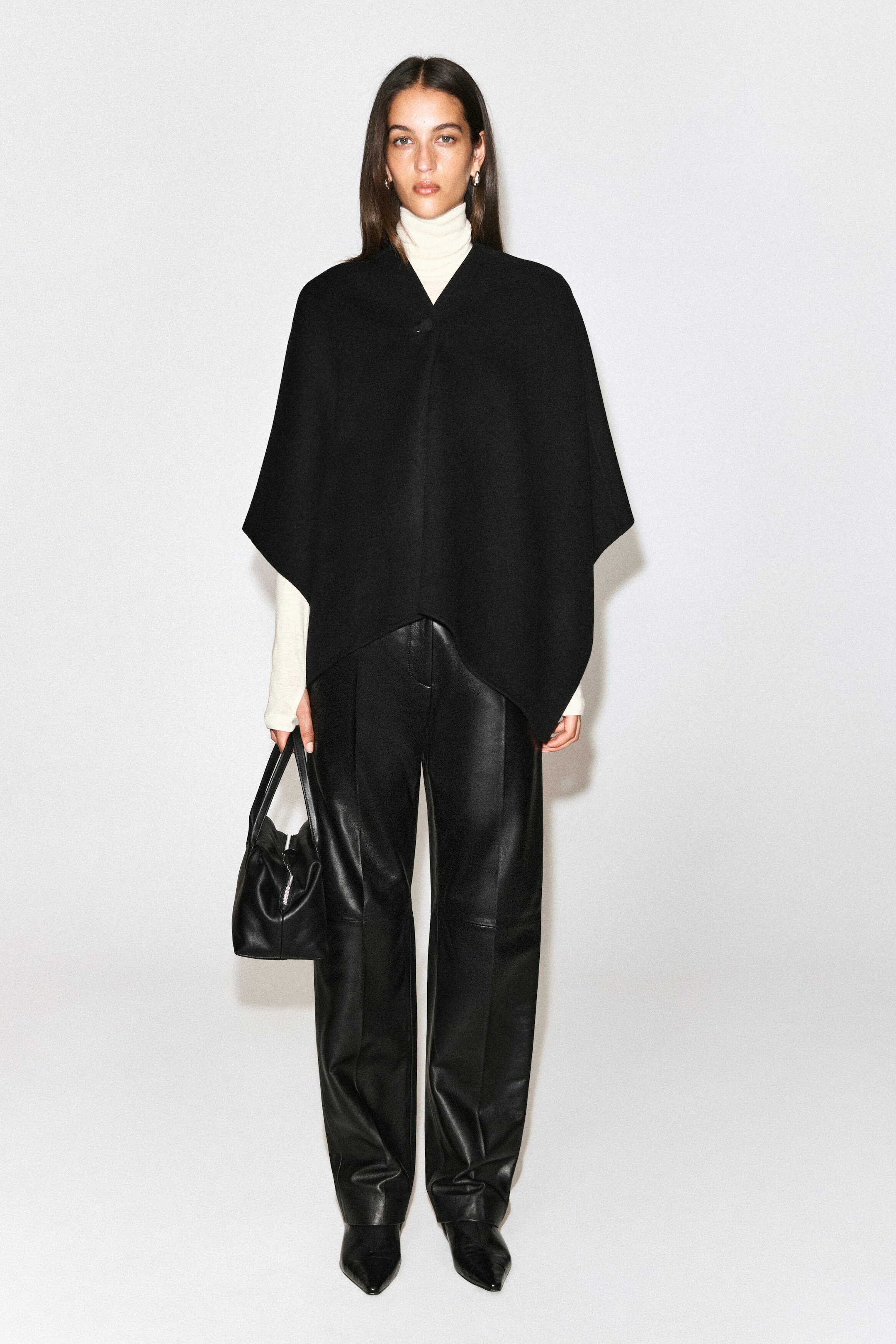 The model is wearing a black double-faced wool-blend cape over a cream-coloured roll-neck sweater and black leather trousers. The cape features a draped silhouette with a V-neckline, a single-button fastening at the collar, and an asymmetrical hemline. The model is wearing a black double-faced wool-blend cape over a cream-coloured roll-neck sweater and black leather trousers. The cape features a draped silhouette with a V-neckline, a single-button fastening at the collar, and an asymmetrical hemline.