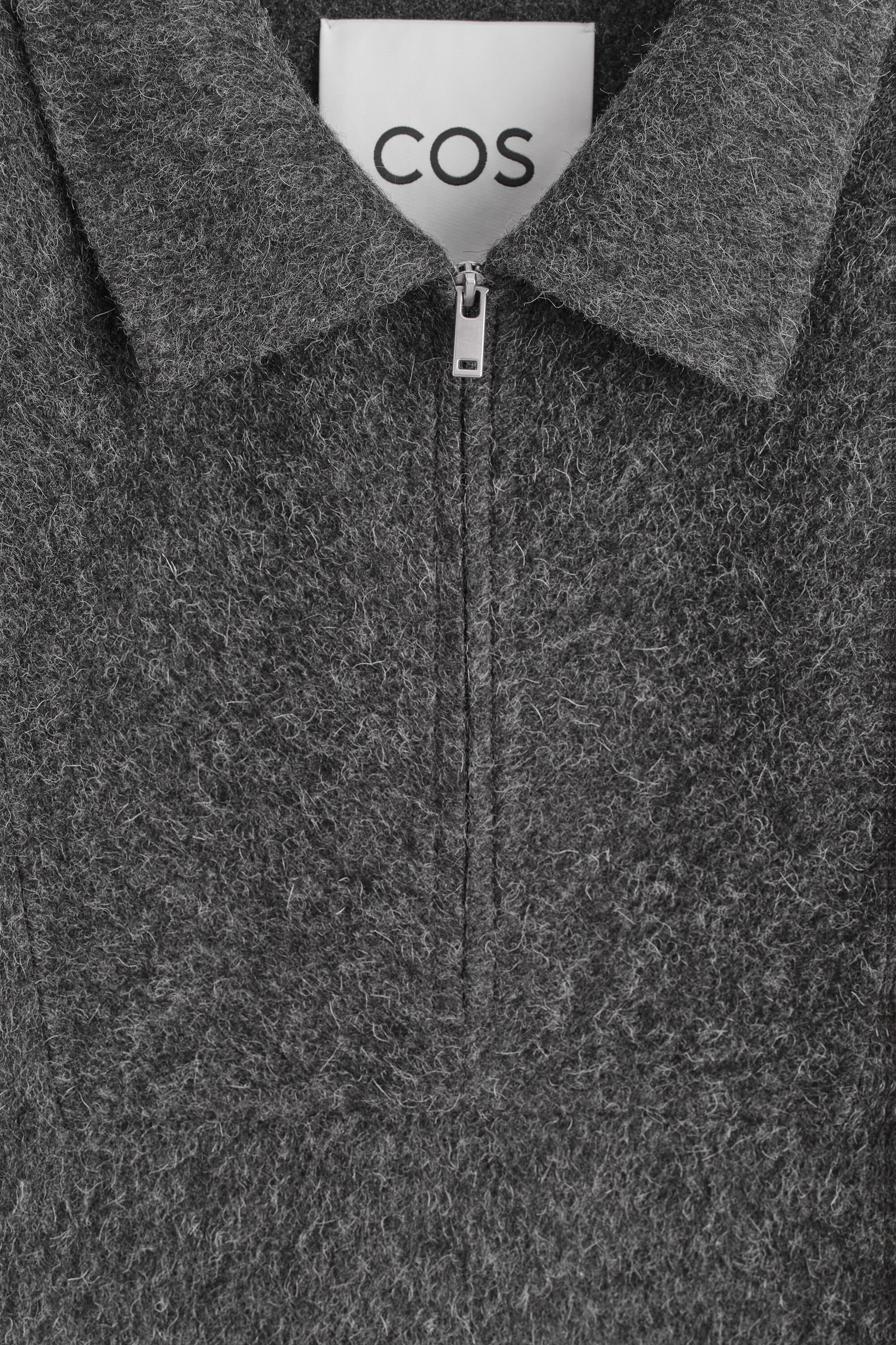 A gray textured garment is displayed in a close-up front view, featuring a pointed collar and a silver-tone half-zip closure. The fabric has a fuzzy, melange-like wool texture, with a white COS brand label visible at the inner neckline.