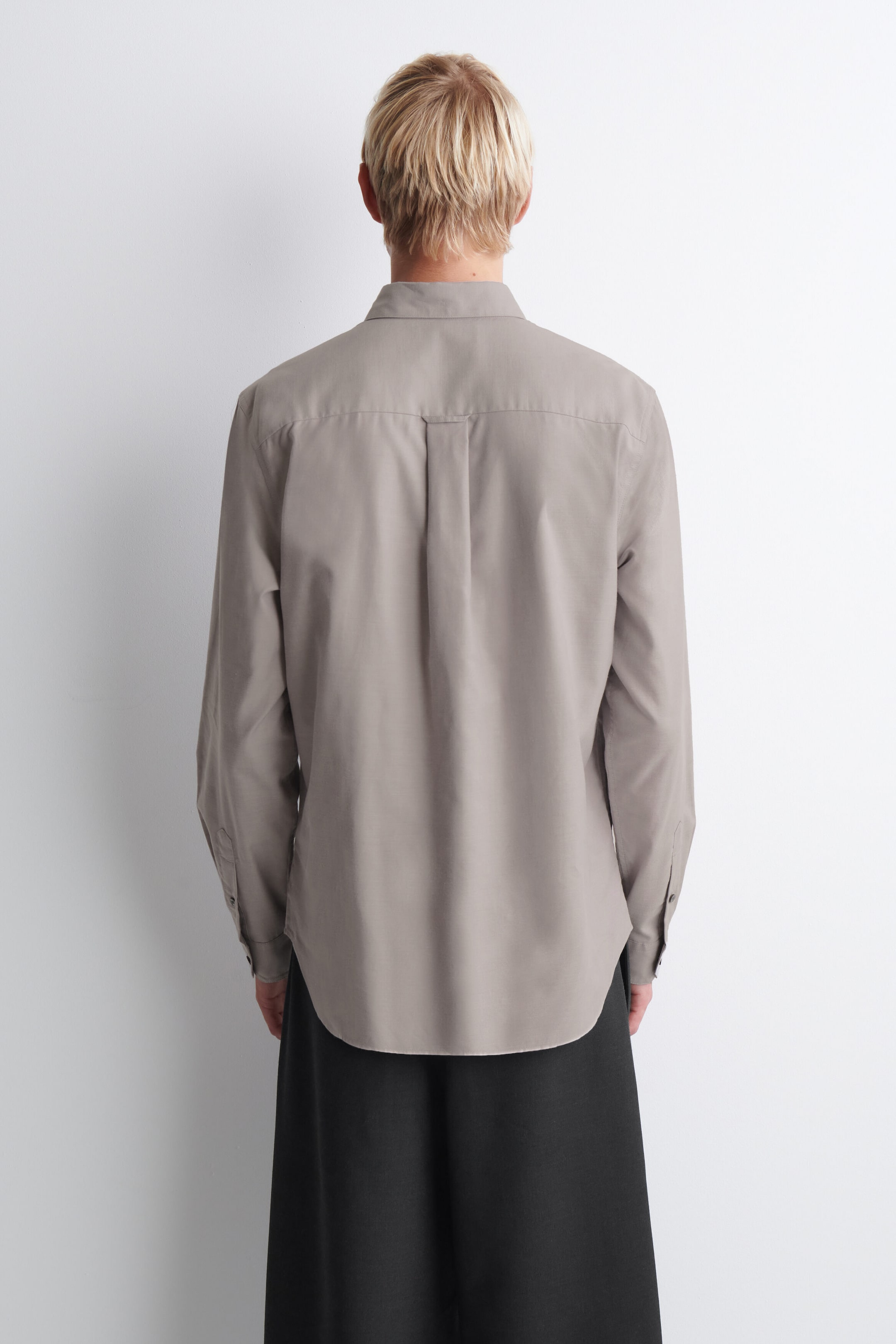 The button-down collar brushed Oxford shirt is shown from the back to highlight its structured design, featuring a horizontal back yoke and a centre box pleat with a locker loop. This long-sleeve muted grey shirt displays a curved hem and buttoned cuffs while draping loosely to showcase its relaxed fit.