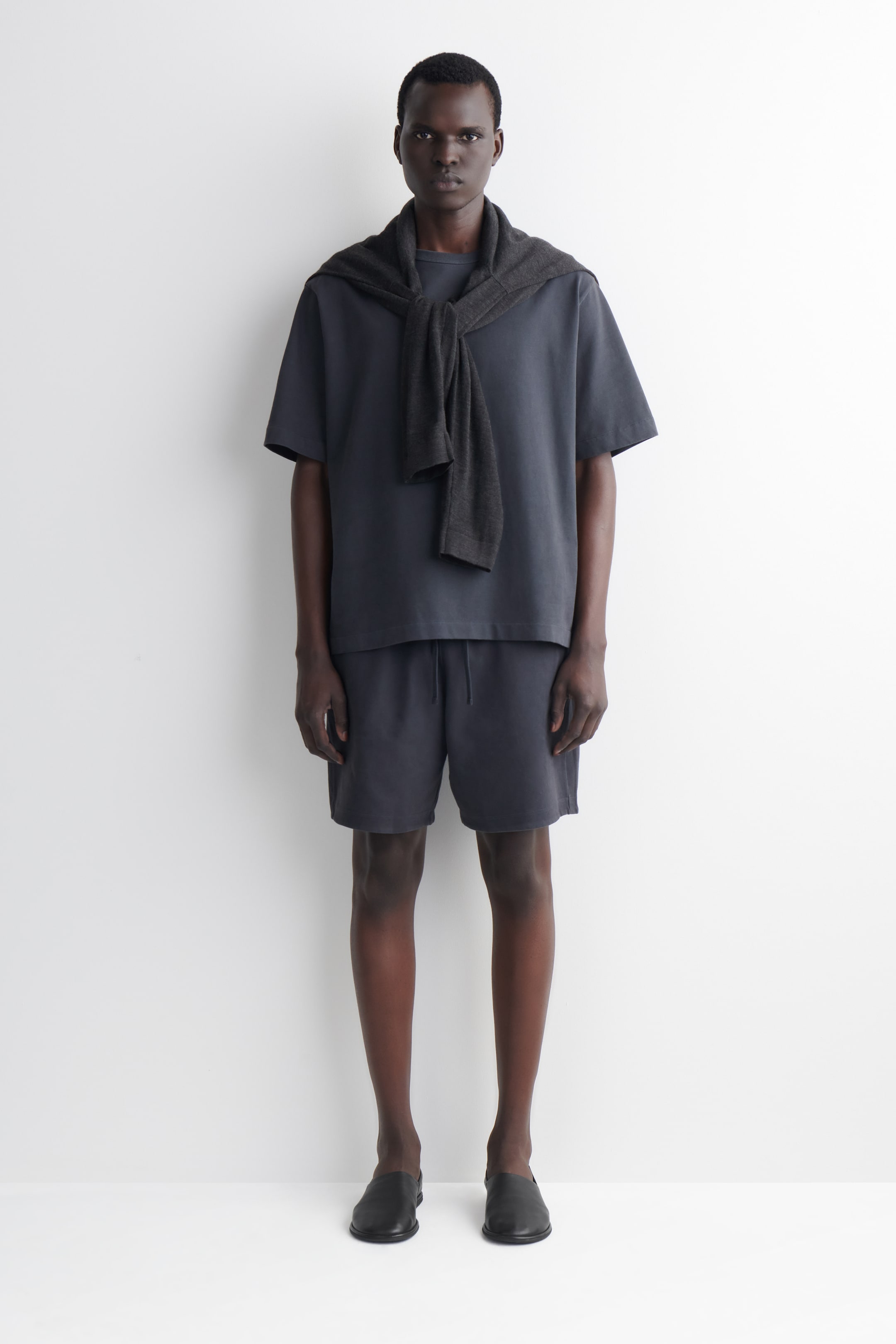 Visualizza immagine più grande: Una t-shirt a maniche corte con collo a giro di colore grigio scuro e pantaloncini con coulisse coordinati vengono indossati, presentando una vestibilità rilassata. Un maglione grigio scuro è drappeggiato sulle spalle e annodato sul davanti, mentre delle scarpe nere slip-on vengono indossate ai piedi.