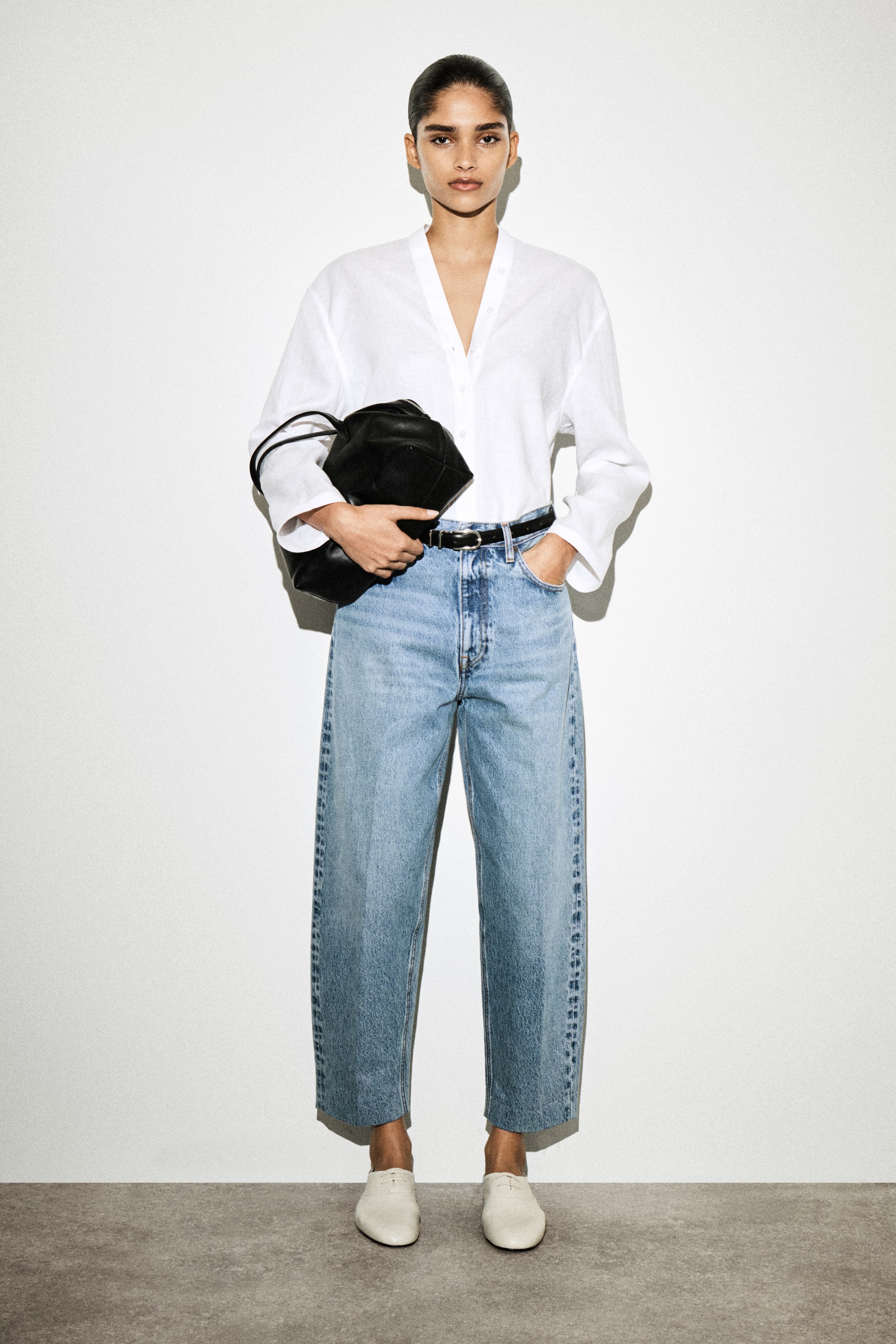 הצגת תמונה גדולה יותר: חולצה לבנה עם שרוולים ארוכים ומחשוף V נלבשת, תחובה בתוך ג'ינס רחב בגזרה קצרה בצבע בהיר, שמוחזק על ידי חגורה שחורה. תיק שחור מקומט מוחזק ביד, ונעליים שטוחות לבנות משלימות את ההופעה.