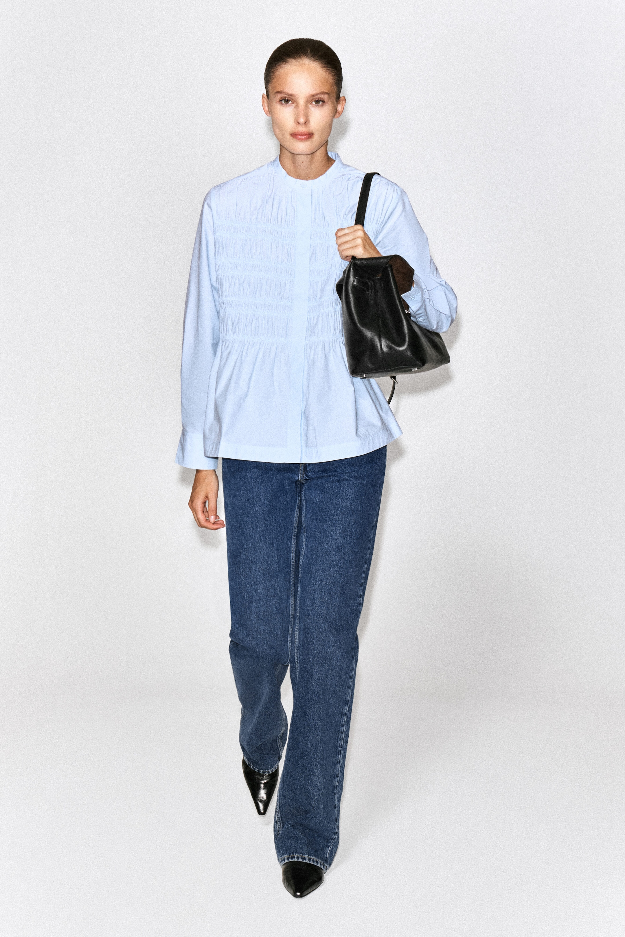 A light blue ruched cotton blouse featuring a band collar, long sleeves, and a smocked chest panel is being worn by the model. The blouse is styled with dark blue jeans and a black leather shoulder bag. A light blue ruched cotton blouse featuring a band collar, long sleeves, and a smocked chest panel is being worn by the model. The blouse is styled with dark blue jeans and a black leather shoulder bag.