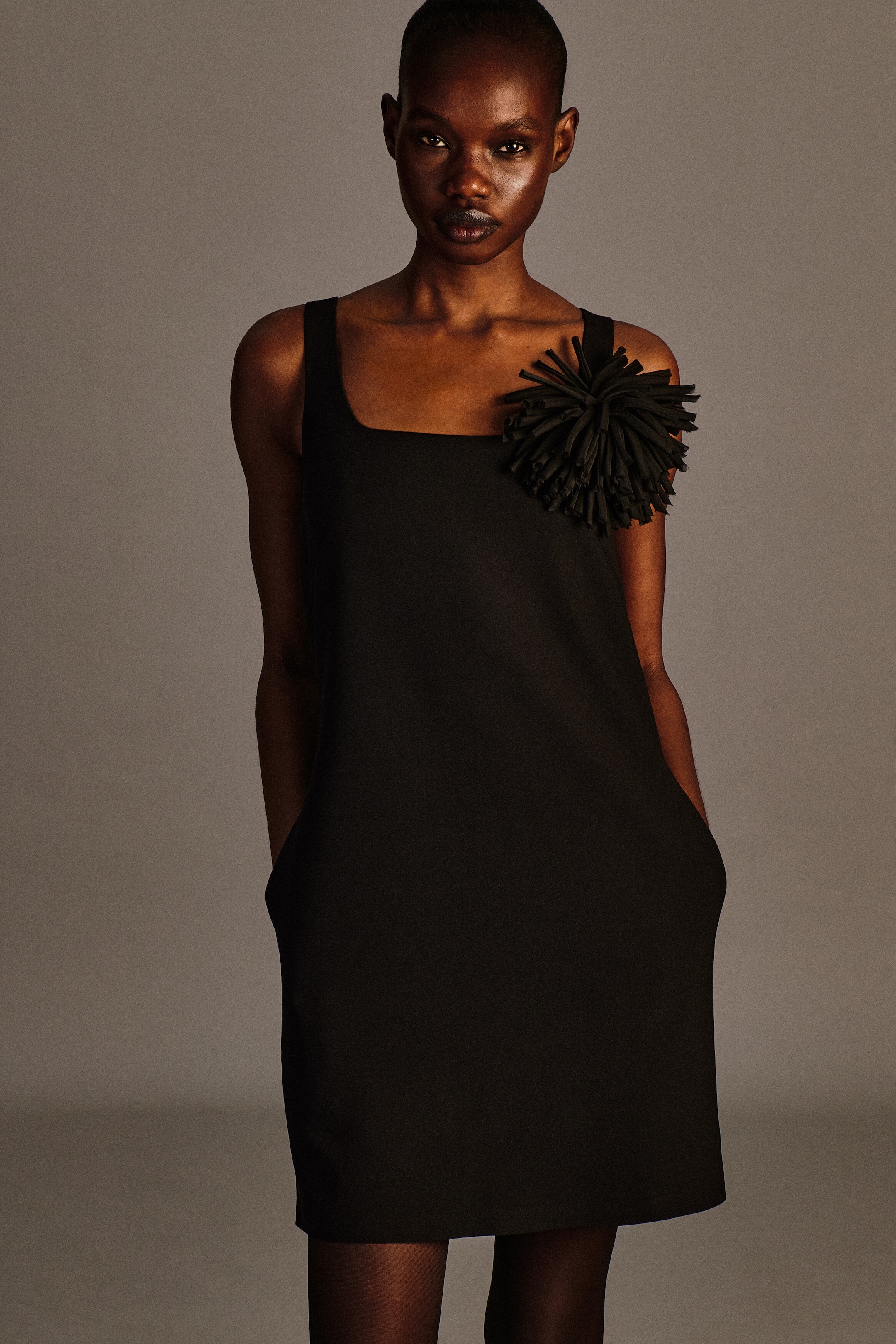 The black square-neck crepe jersey dress is displayed from the front in a sleeveless, relaxed silhouette. A large textured black floral rosette brooch accents the left shoulder strap, adding dimensional detail to the clean design. The black square-neck crepe jersey dress is displayed from the front in a sleeveless, relaxed silhouette. A large textured black floral rosette brooch accents the left shoulder strap, adding dimensional detail to the clean design.