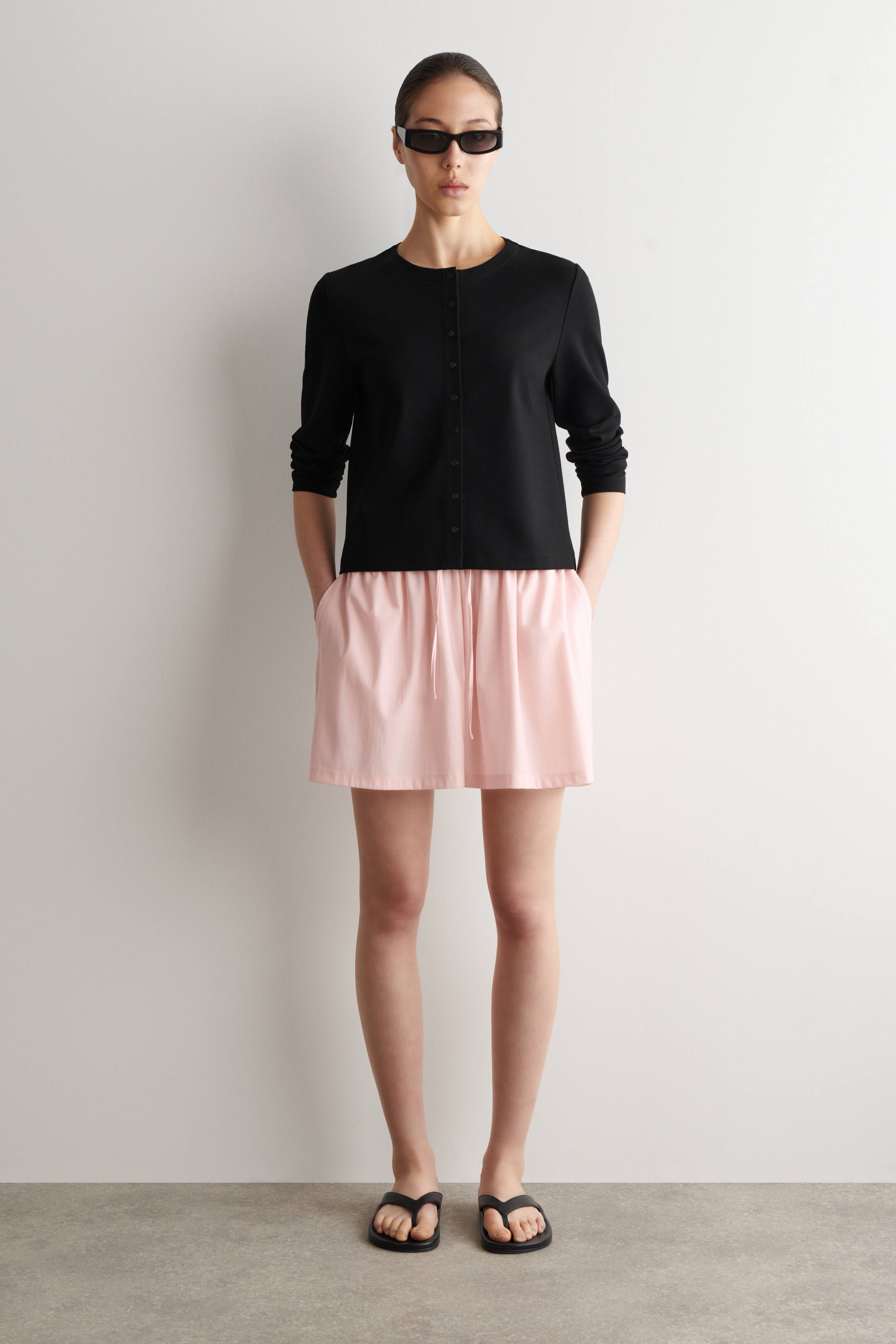 Se større billede: Lyserøde shorts med en rynket talje og tonale snoretræk bæres af modellen. Modellen har hænderne i shortsens lommer og styler dem med en sort langærmet top.