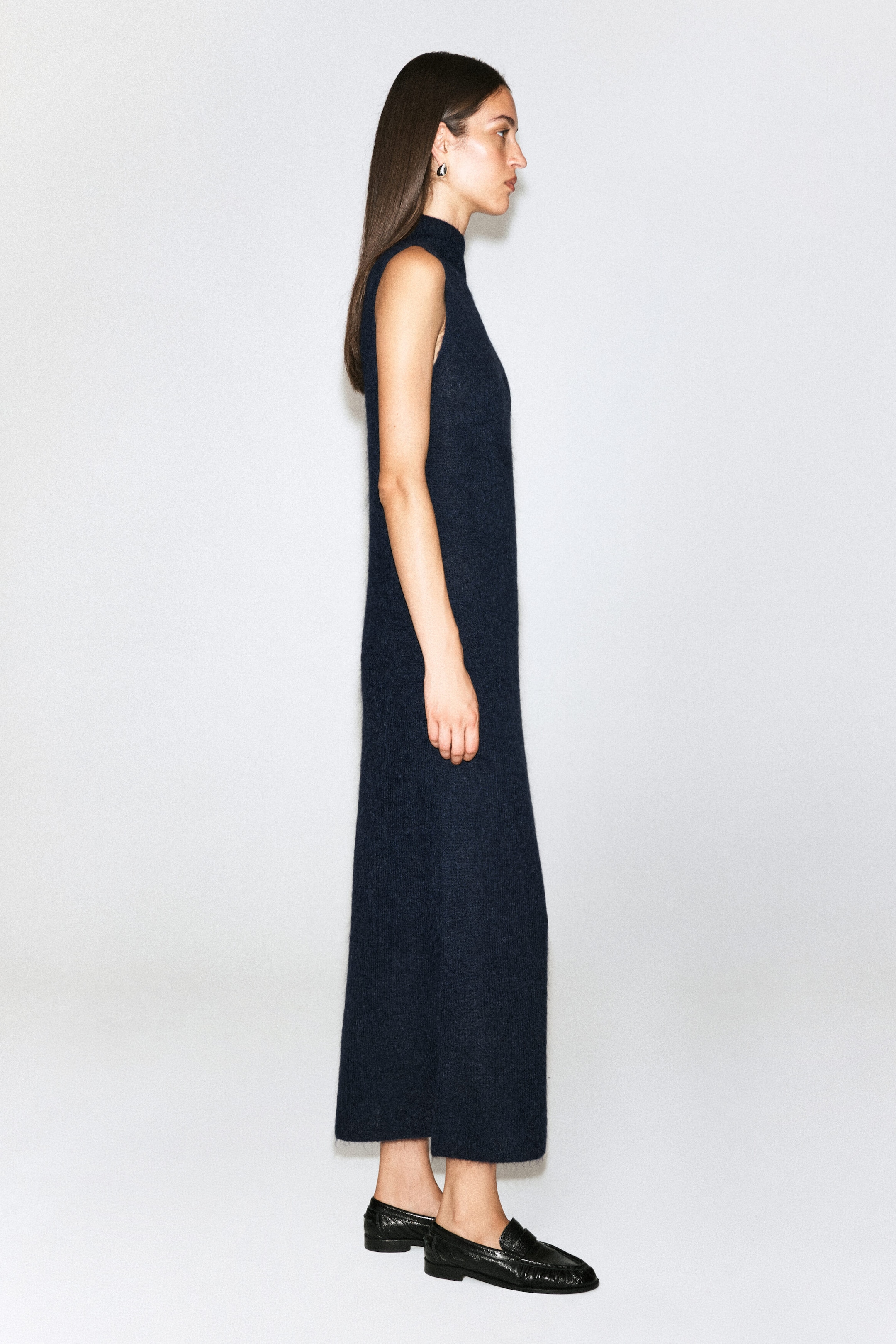 A brushed funnel-neck dress in dark navy is worn by the model, shown in side profile. The sleeveless, ankle-length dress features a straight silhouette and a soft textured finish, styled with flat black leather loafers.